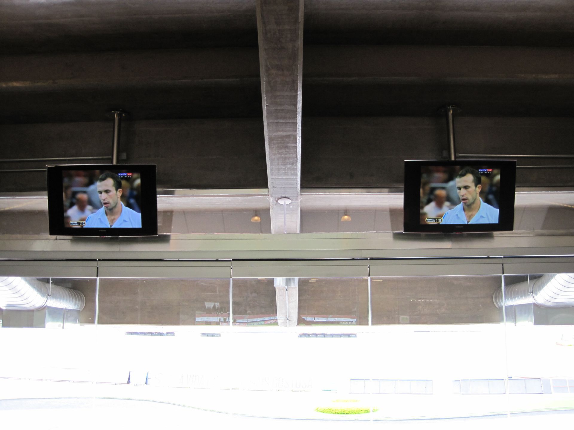 Duas TVs instaladas no teto de um estádio exibem o mesmo homem conversando com uma mulher.
