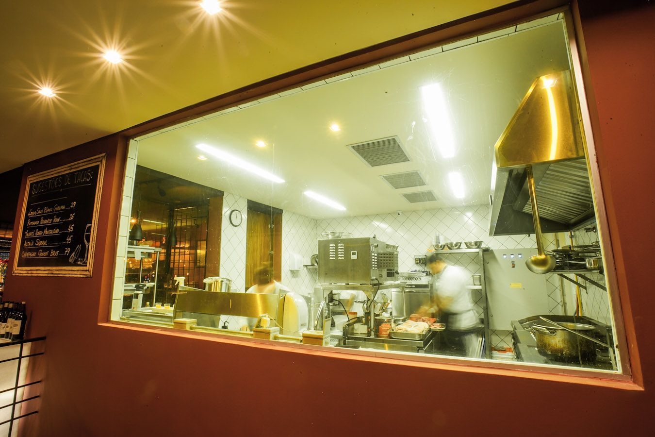 Vista noturna da cozinha de um café com iluminação aconchegante através de uma grande janela.