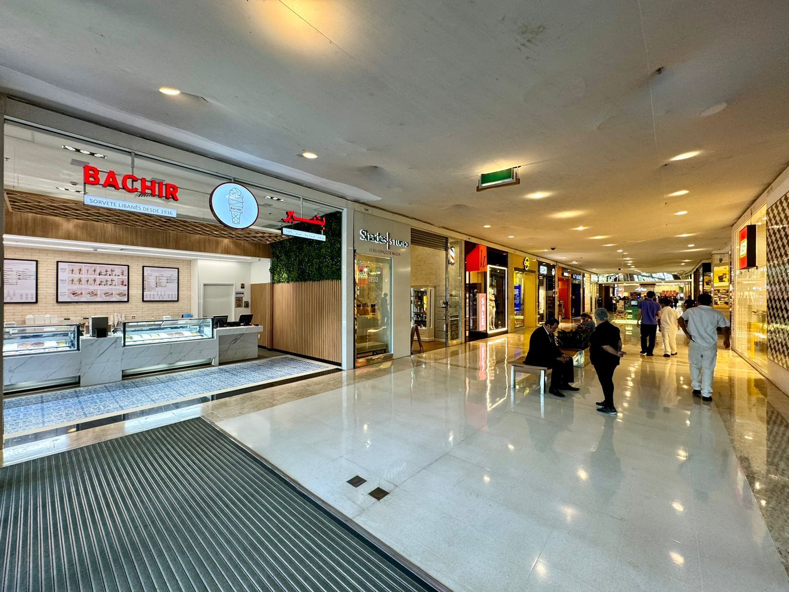 Corredor iluminado de um shopping center com um balcão de padaria à esquerda e compradores circulando pelo hall.