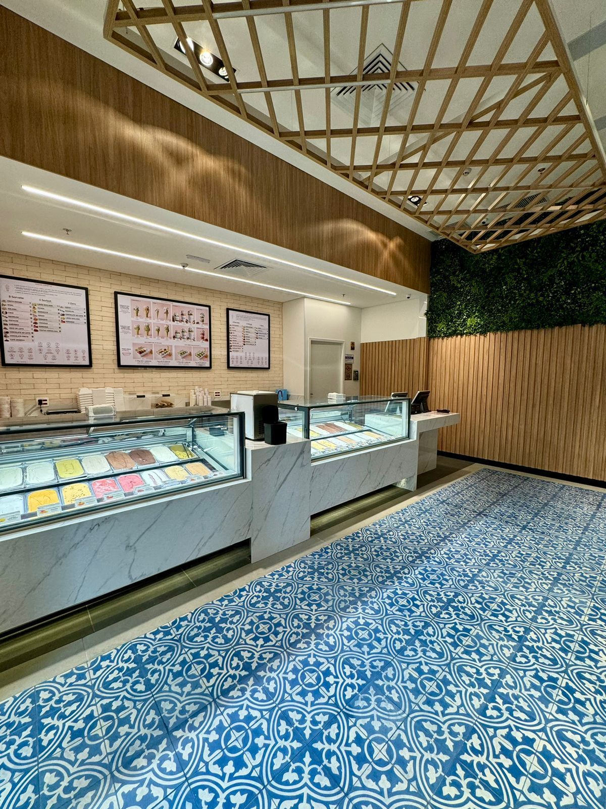 Interior moderno de sorveteria com vitrines de vidro, detalhes em madeira e piso de ladrilho azul estampado.