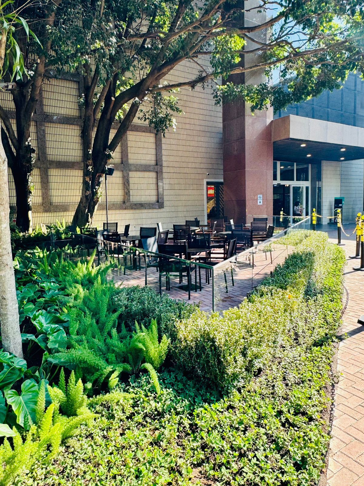 Mesas de café ao ar livre ao lado de um prédio bege, com plantas e árvores verdejantes em primeiro plano.