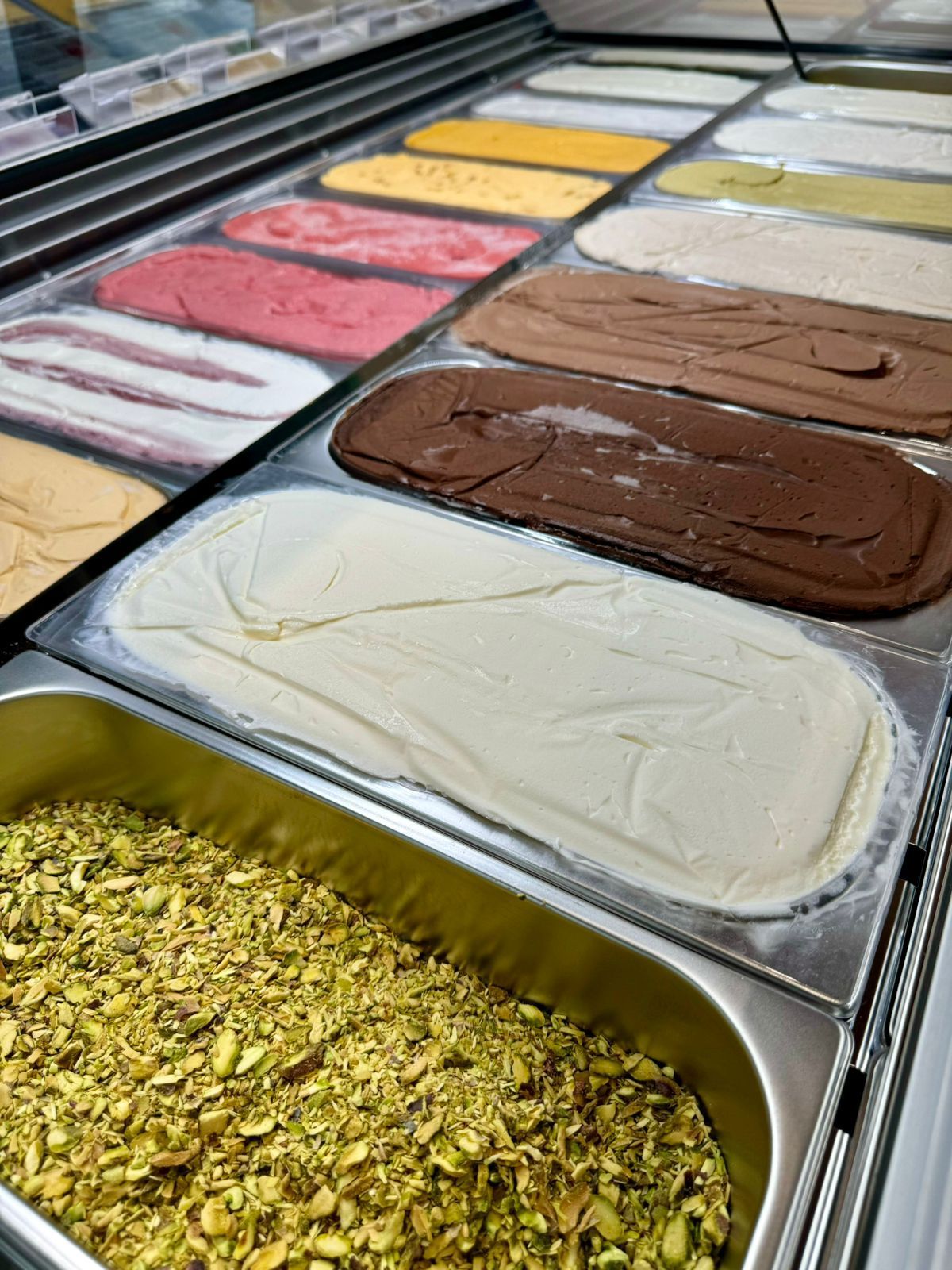 Variedade de sabores de gelato em bandejas de aço inoxidável em uma sorveteria, incluindo chocolate, baunilha e pistache.