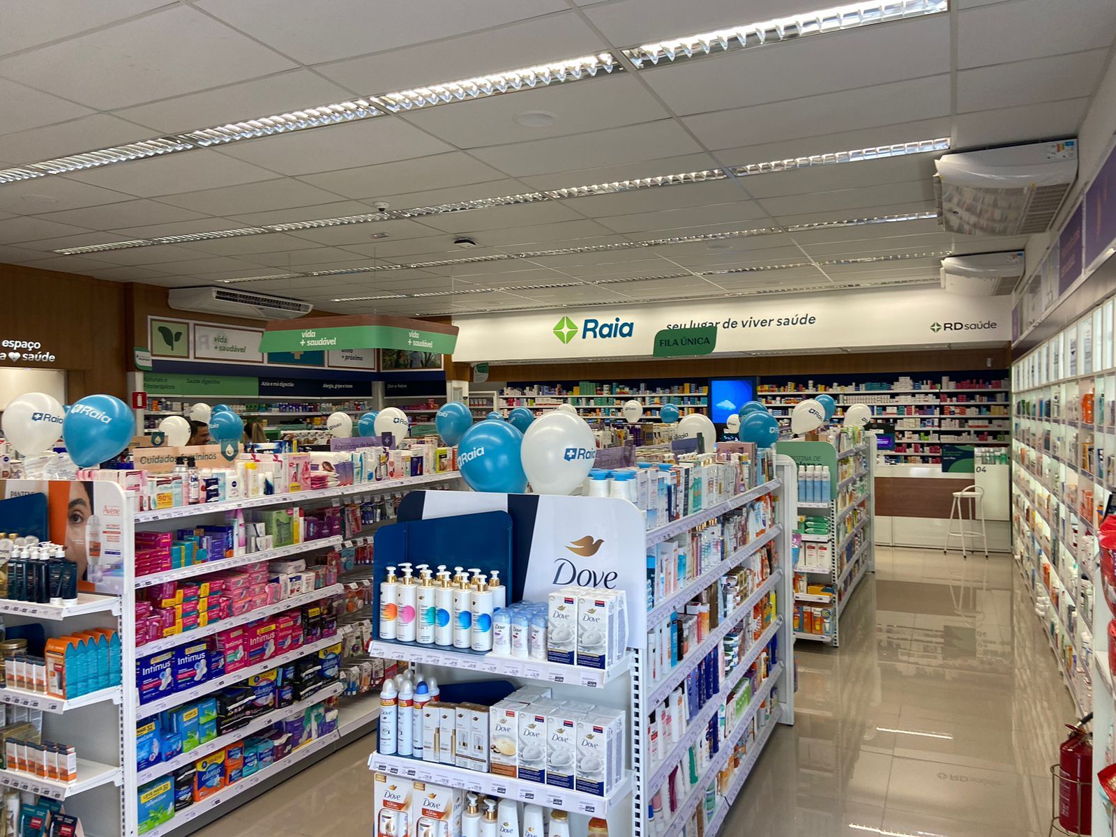 Corredor de farmácia com prateleiras de medicamentos e produtos de higiene pessoal, balões azuis e iluminação forte no teto.