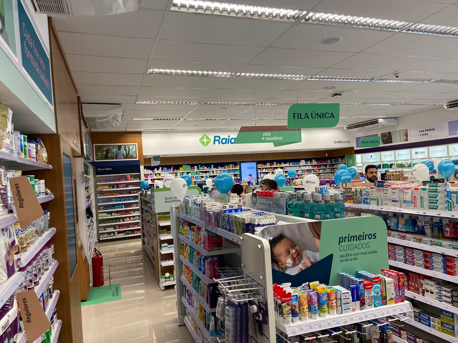 Corredor de farmácia bem iluminado, com prateleiras abastecidas, placas penduradas e um cliente olhando os produtos na loja.
