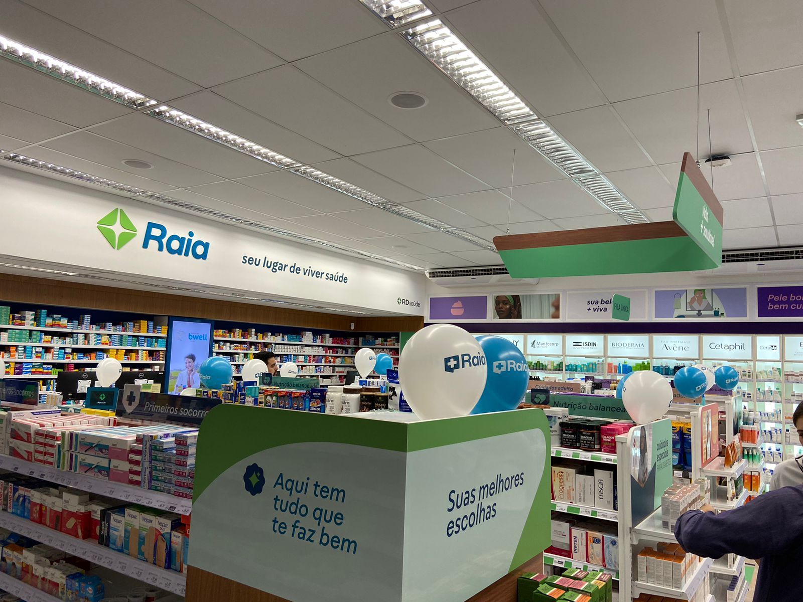 Corredor da farmácia com placa da Raia, prateleiras abastecidas e um cliente próximo ao balcão.