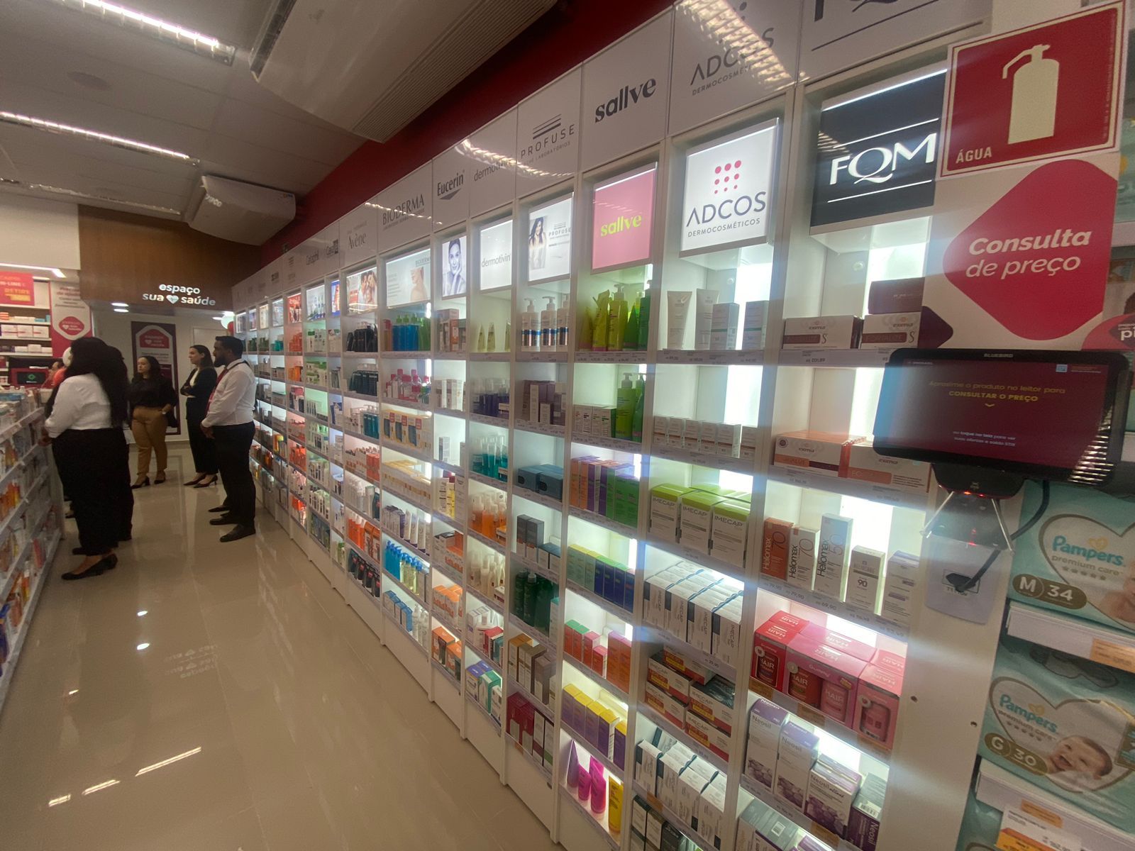 Corredor da farmácia com prateleiras iluminadas de medicamentos e alguns clientes olhando os produtos.