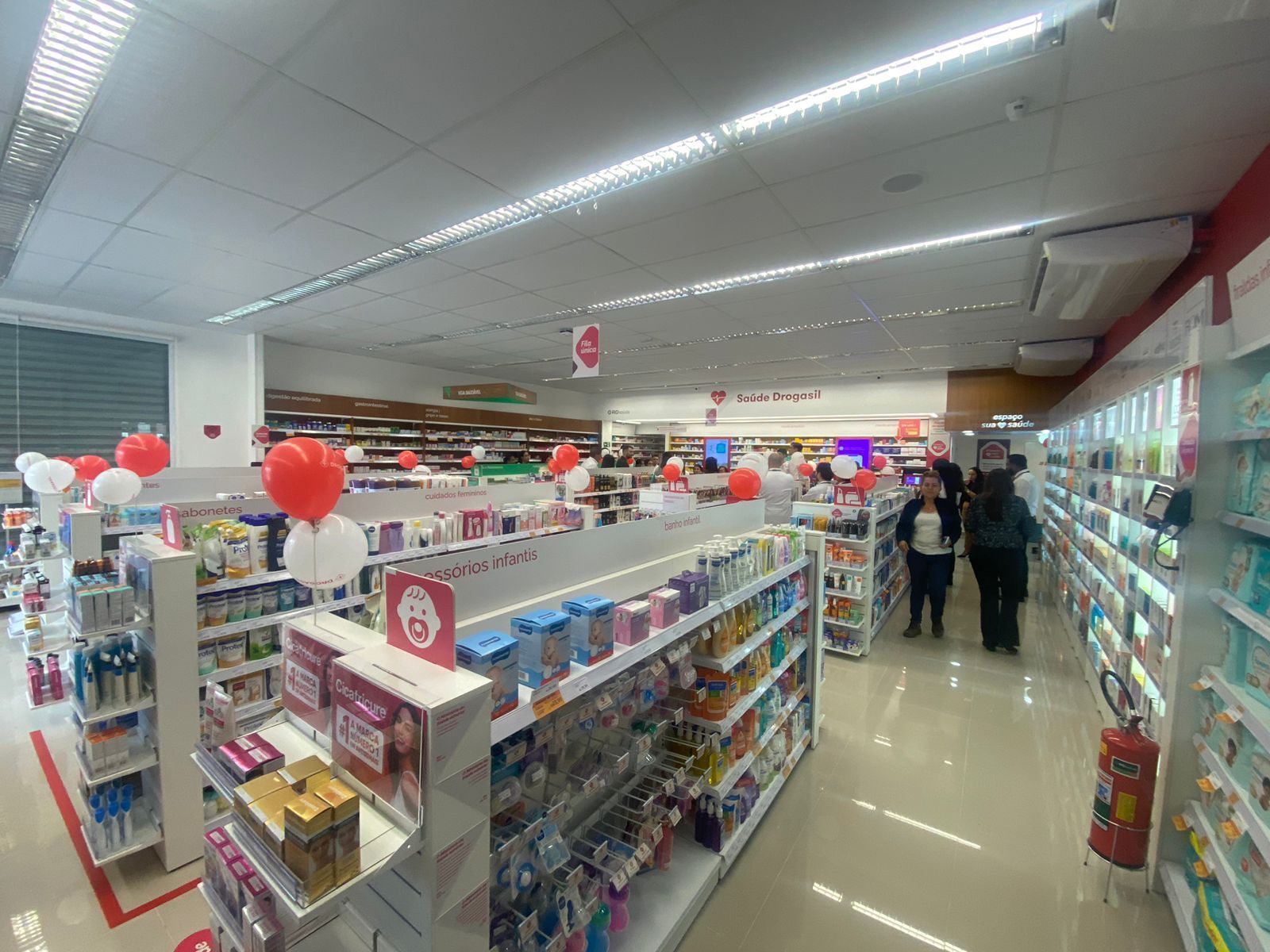 Corredor de farmácia iluminado com prateleiras de medicamentos, balões vermelhos e dois clientes entrando.