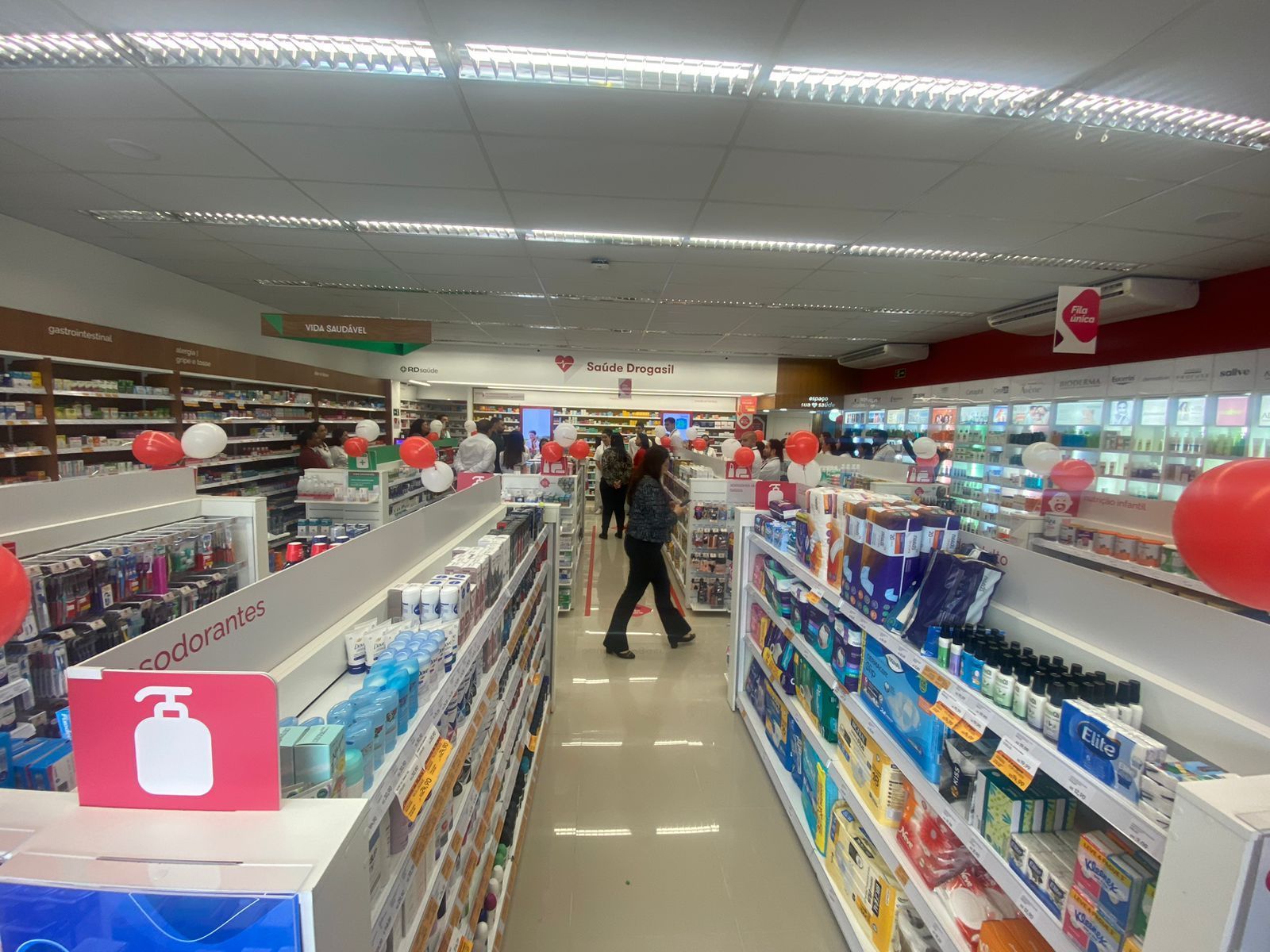Um corredor de loja com prateleiras de artigos de higiene pessoal e produtos de limpeza, com uma pessoa em pé no centro.