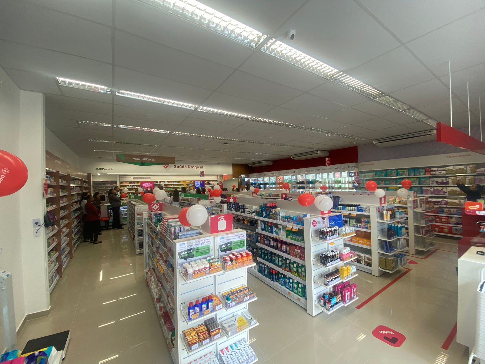Interior luminoso de uma loja de conveniência com prateleiras de salgadinhos e bebidas, balões vermelhos e brancos e alguns clientes dentro.