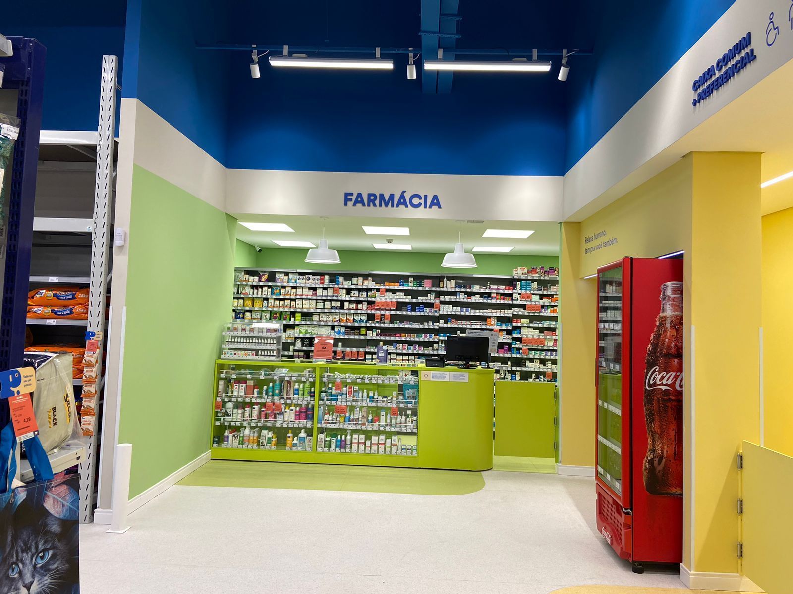 Interior de farmácia com paredes verdes, prateleiras de medicamentos e uma máquina de venda automática vermelha.