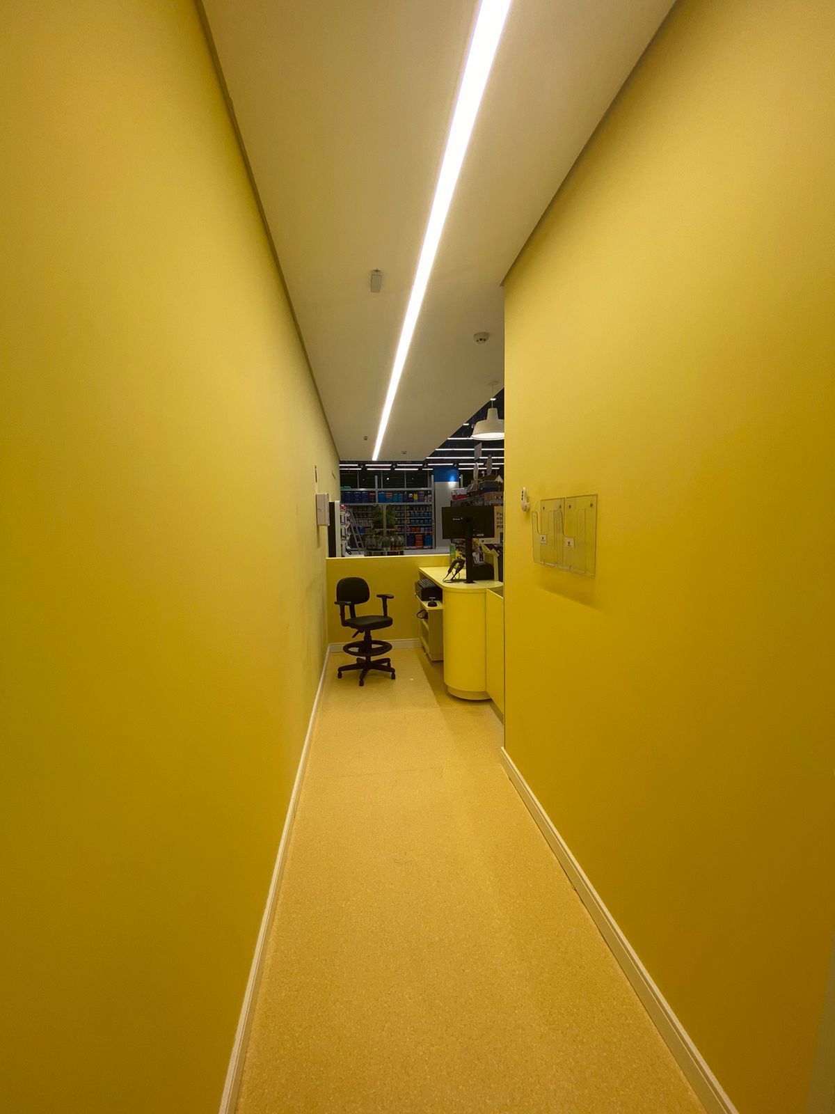 Corredor estreito e amarelo com uma cadeira e uma área de escritório no final, iluminado por uma lâmpada fluorescente no teto.
