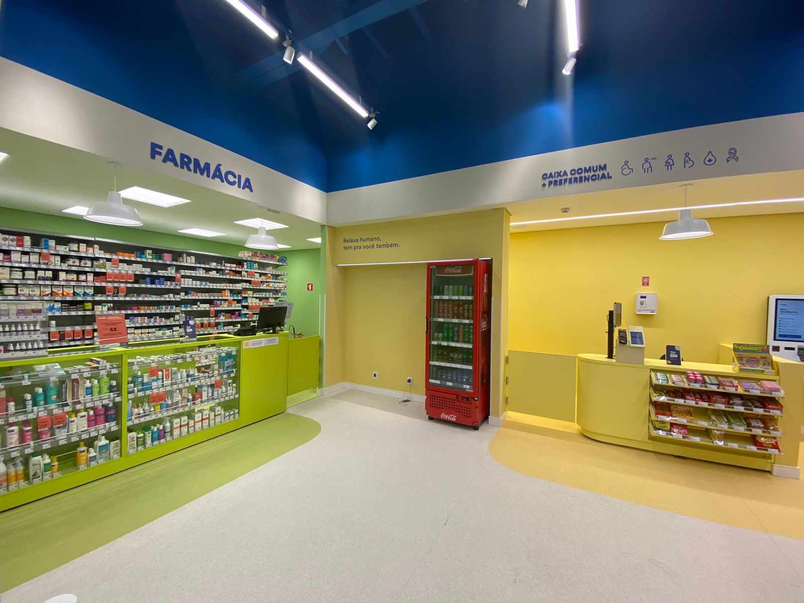 Interior de farmácia vazio com paredes verdes e amarelas, prateleiras de produtos e um refrigerador vermelho.
