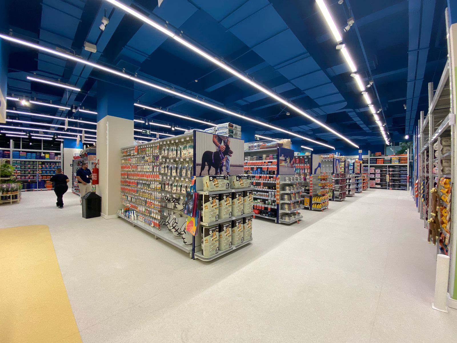 Corredor de supermercado bem iluminado com prateleiras, luzes azuis no teto e alguns clientes ao longe.