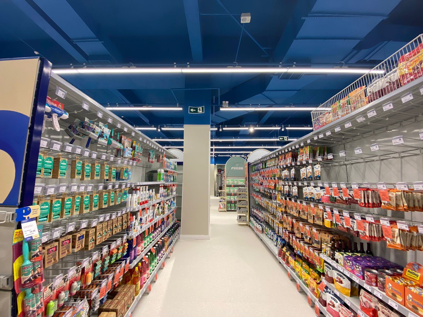 Corredor de supermercado iluminado com prateleiras de produtos coloridos embalados sob luzes azuis no teto.