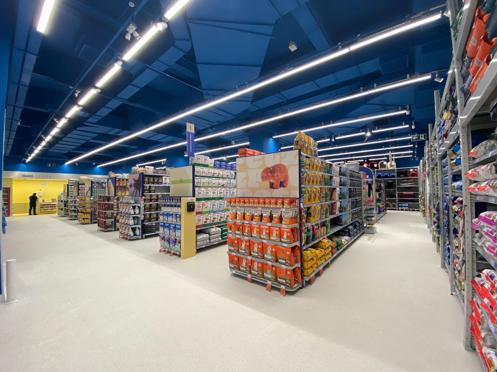 Amplo corredor de supermercado com prateleiras de produtos embalados sob luzes azuis brilhantes no teto.