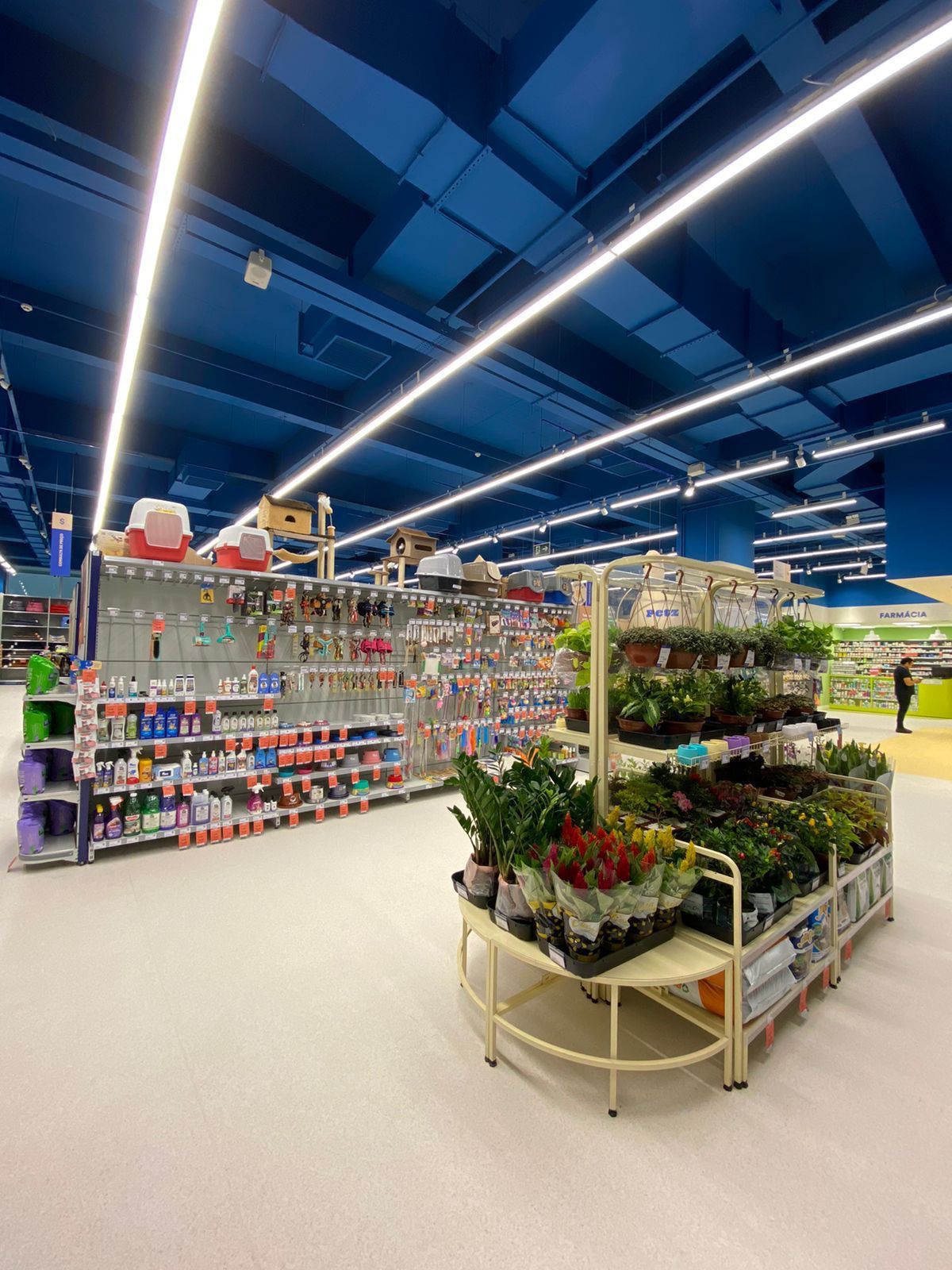 Corredor iluminado de um centro de jardinagem com plantas em vasos, prateleiras de suprimentos e luzes azuis no teto.