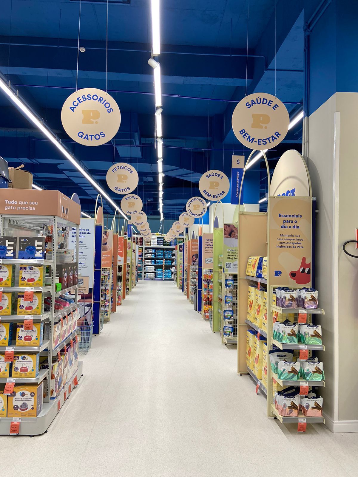 Corredor em um supermercado bem iluminado, com prateleiras de lanches e letreiros azuis no teto.