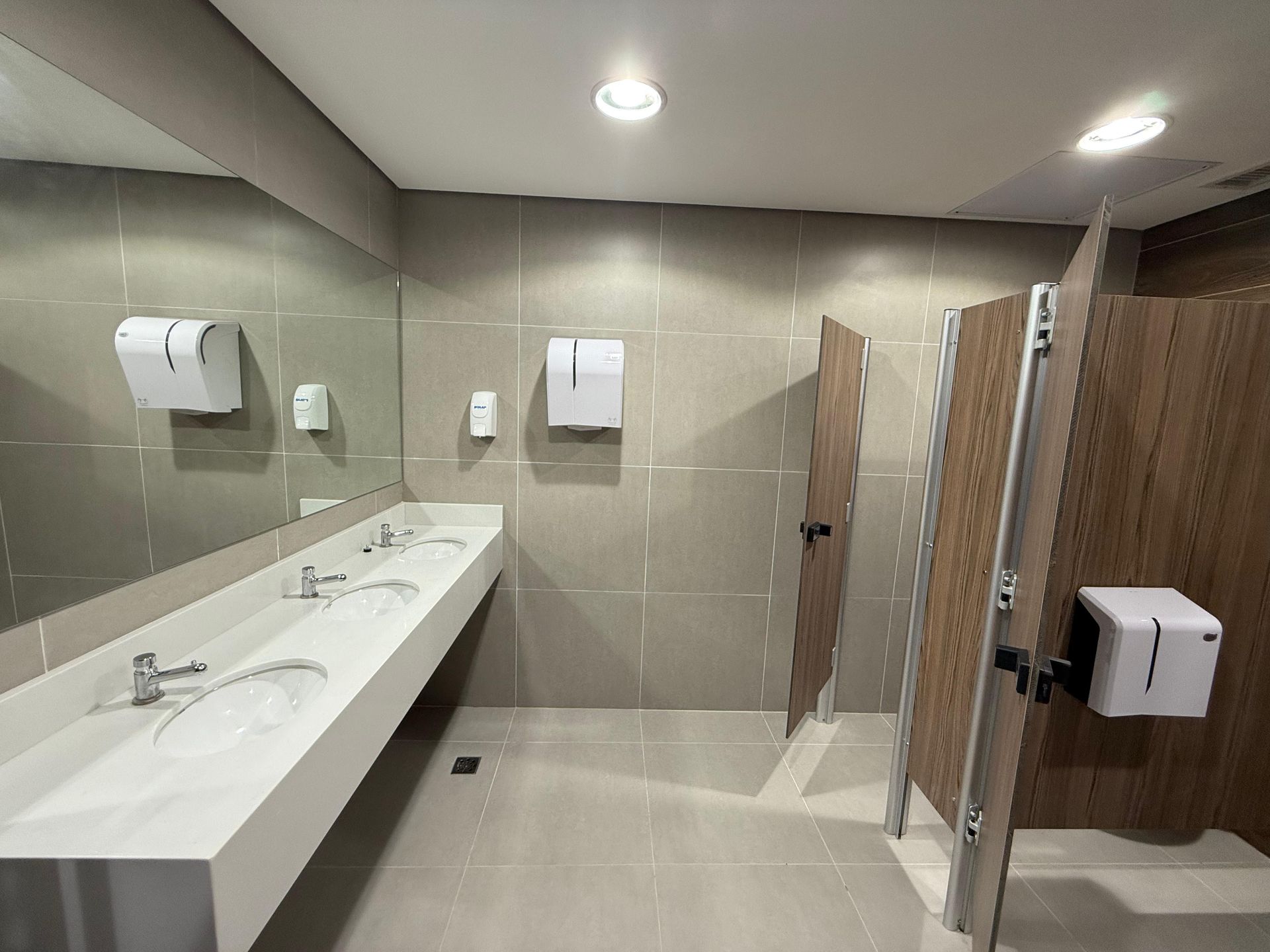 Banheiro moderno com pias compridas brancas, paredes revestidas com azulejos bege e cabines sanitárias com iluminação embutida.