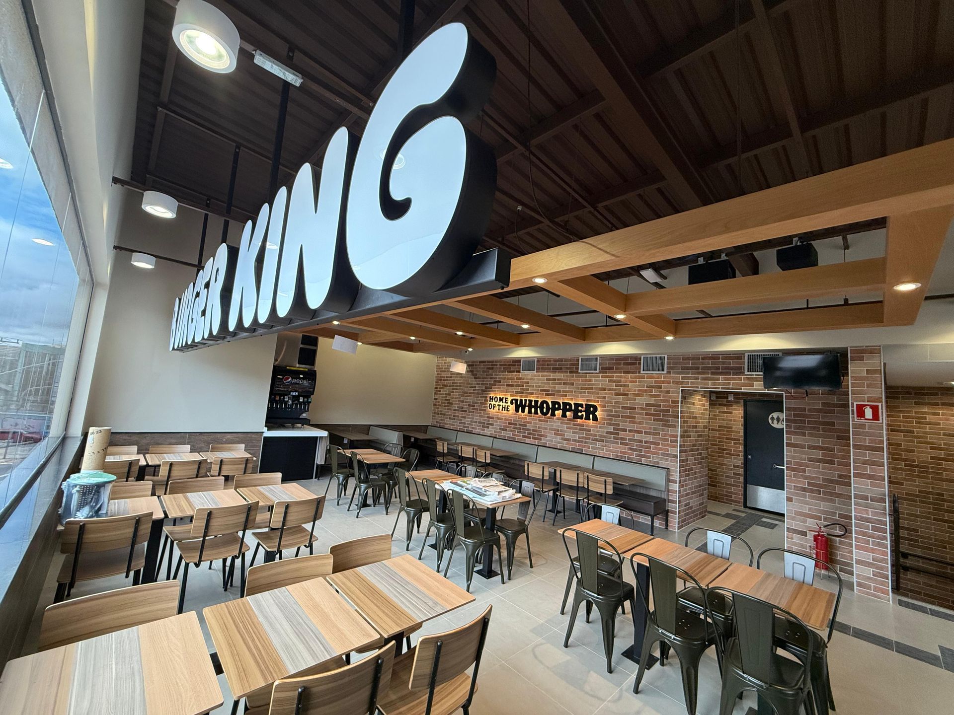 Interior de restaurante moderno com mesas de madeira, paredes de tijolos e grande placa suspensa acima da área de mesas.
