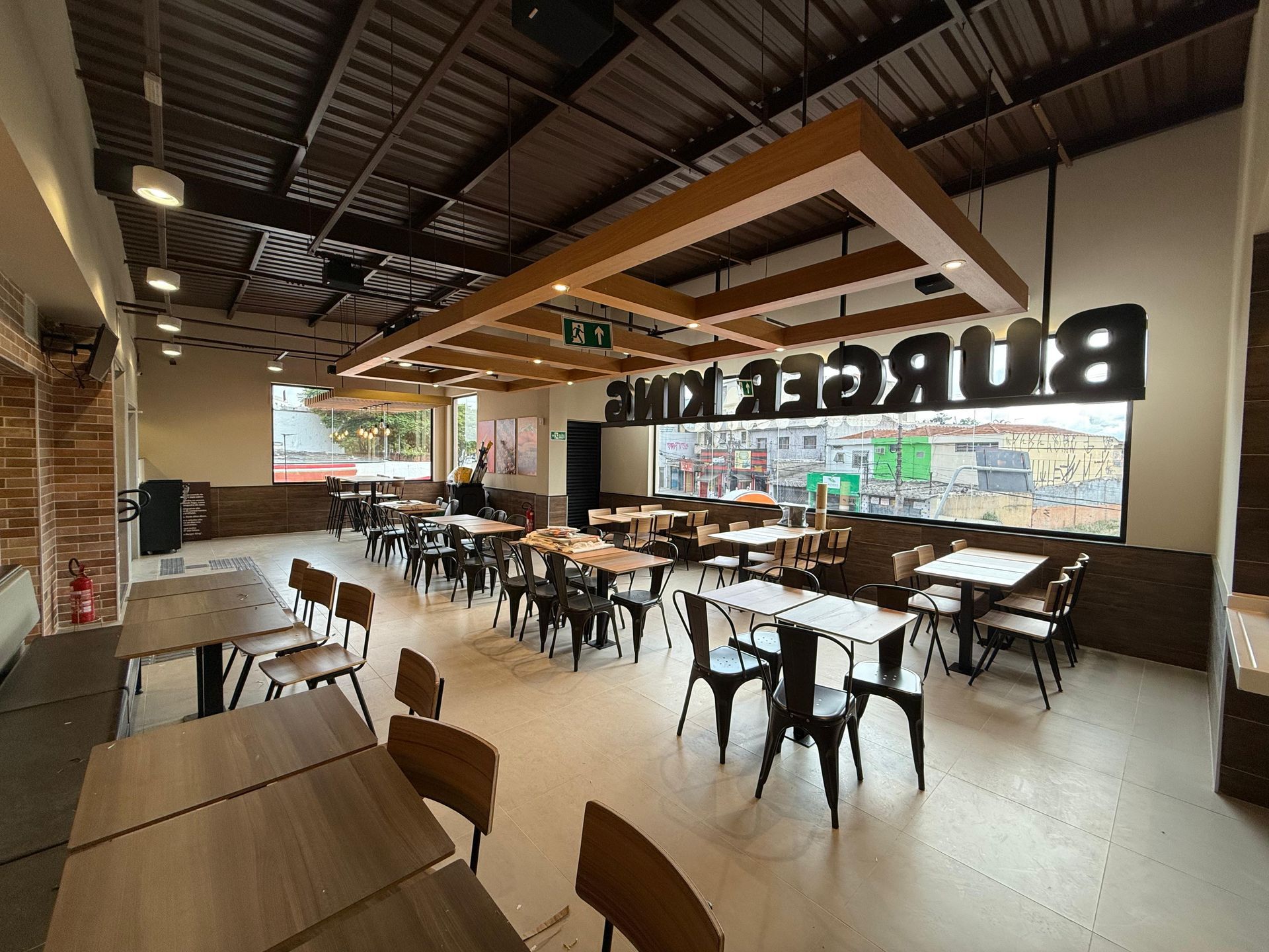 Restaurante moderno e vazio, com mesas de madeira, cadeiras pretas e grandes janelas voltadas para a rua.