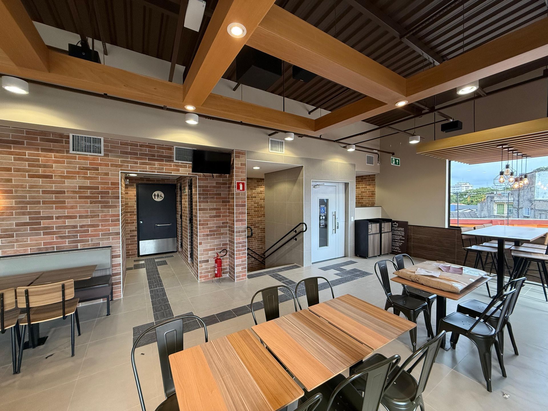 Interior de café moderno com mesas de madeira, cadeiras pretas, paredes de tijolos e grandes janelas com vista para a água.