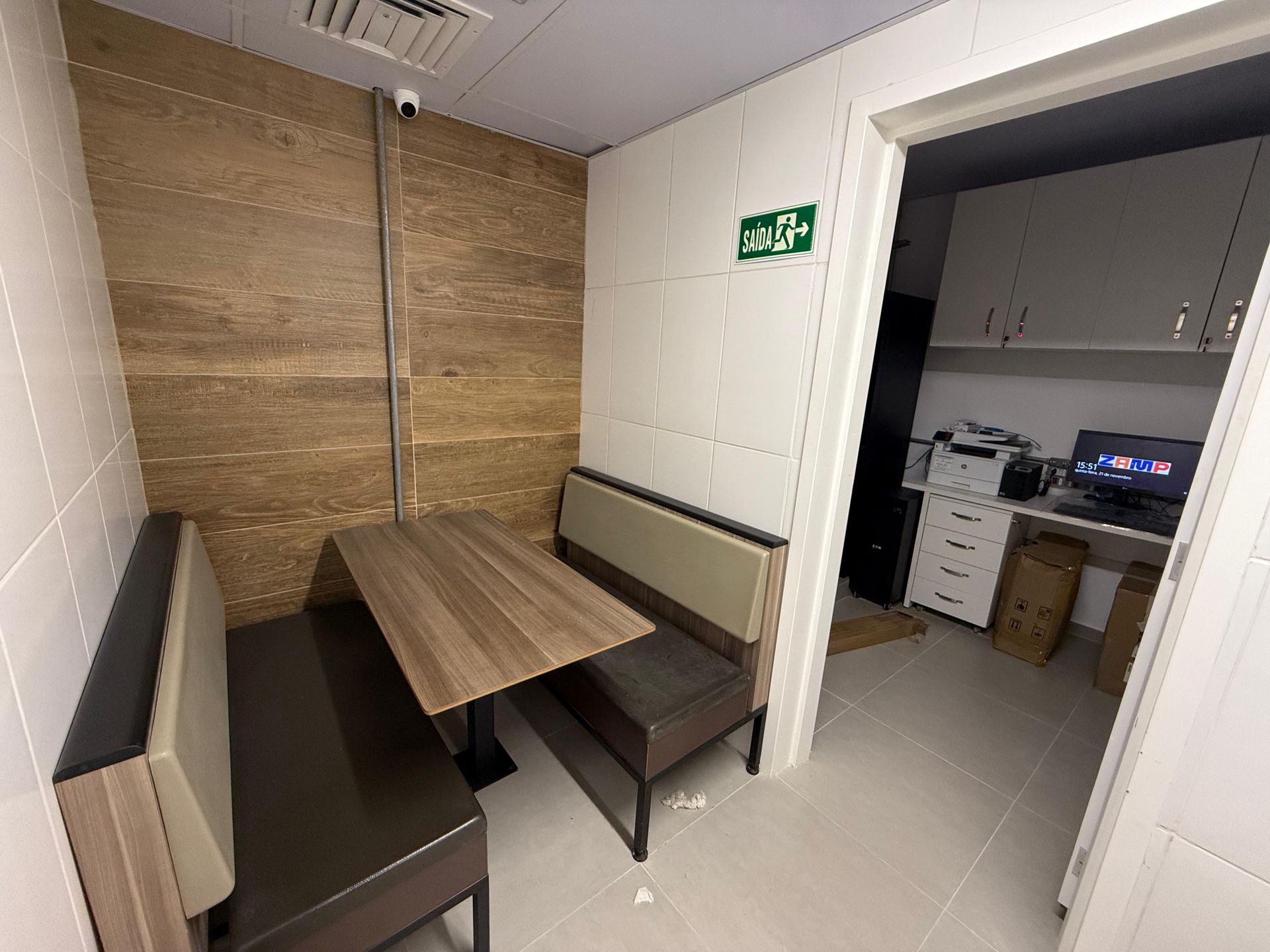 Pequena sala de descanso com mesa de madeira, banco, radiador e porta aberta para uma lavanderia.