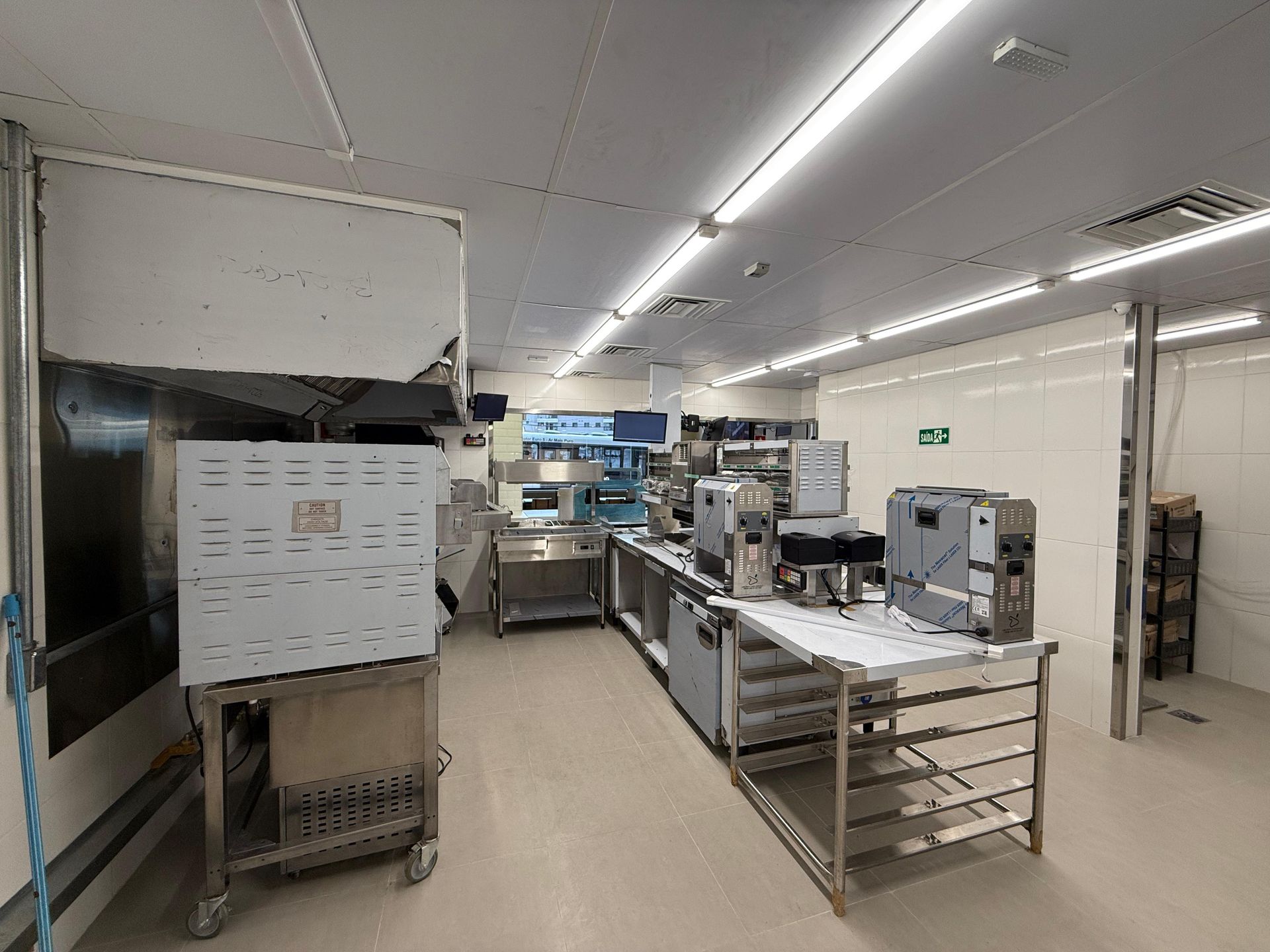 Cozinha industrial com mesas de preparação e equipamentos em aço inoxidável em um ambiente claro com piso de azulejo.
