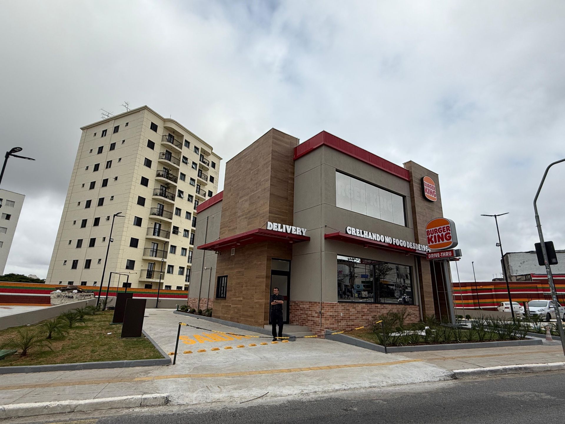 Restaurante de comida rápida na rua, com telhado vermelho e fachada de tijolos, ao lado de um prédio alto de apartamentos.