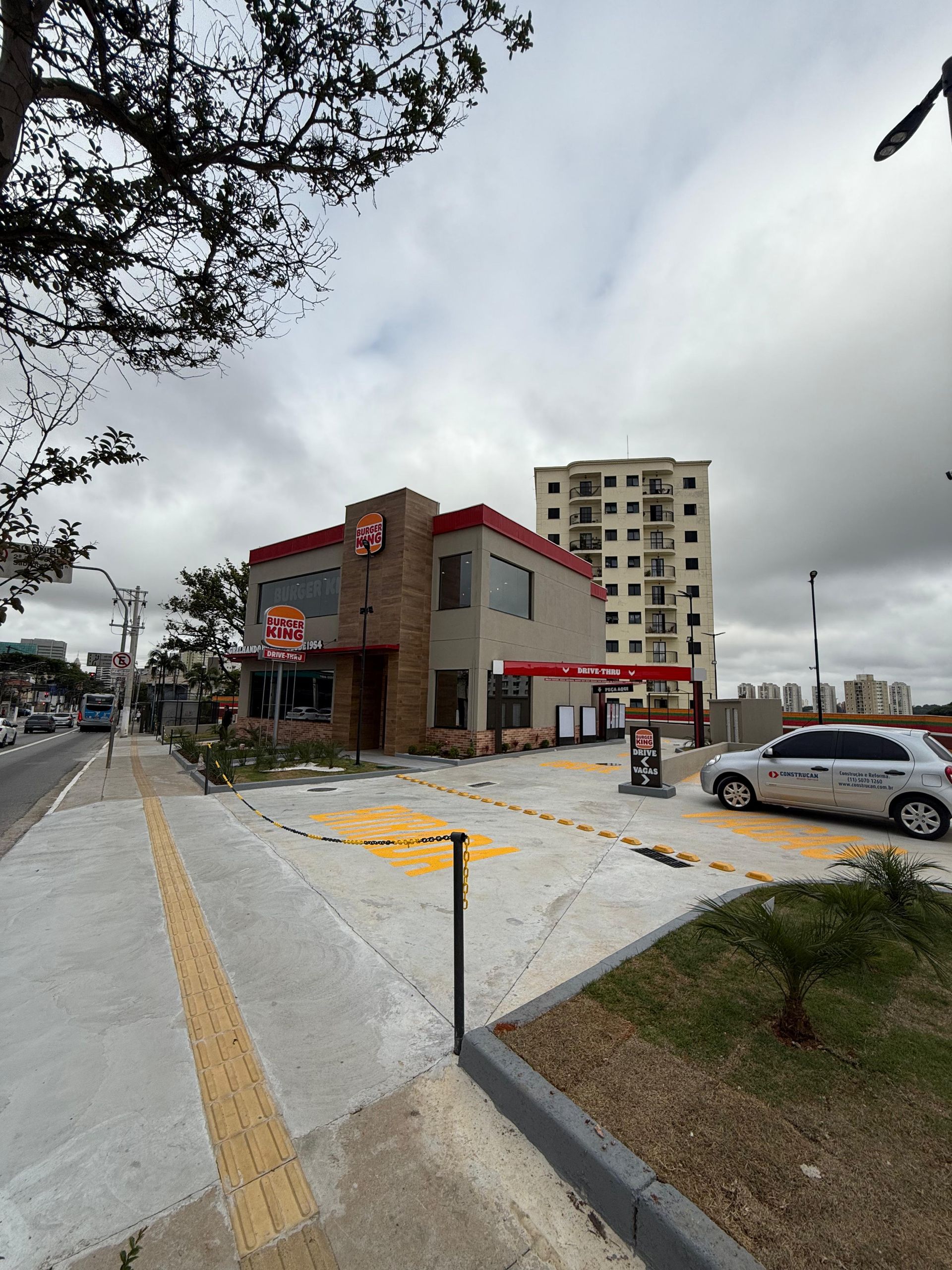 Cafeteria de esquina com mesas ao ar livre junto à calçada e à rua, sob um céu nublado.