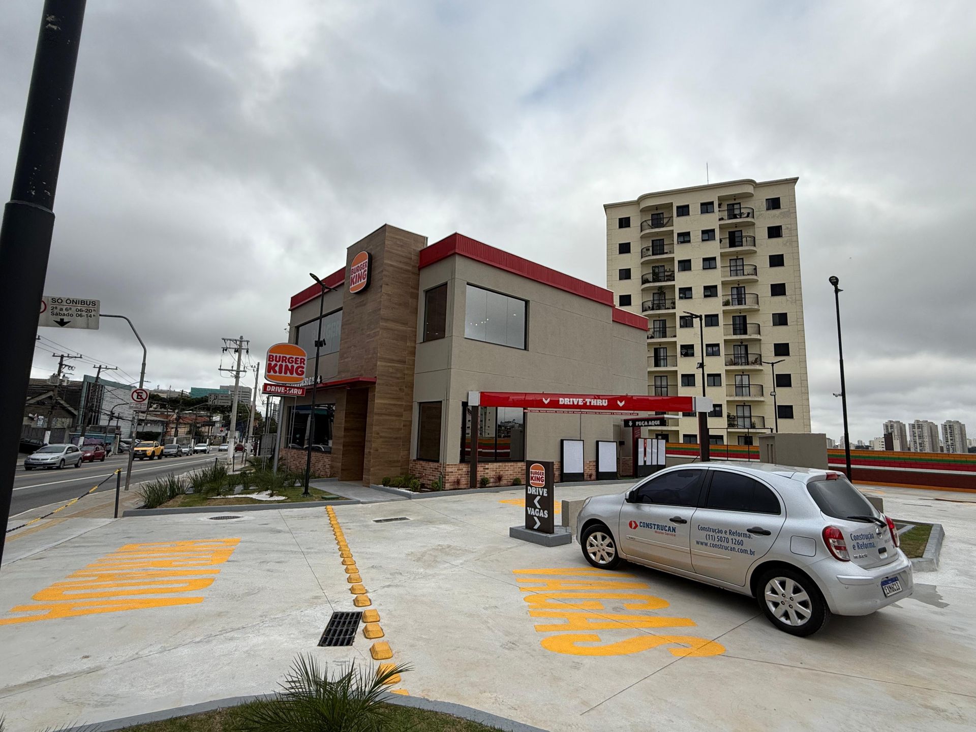 Um drive-thru de fast-food na rua, com um carro branco estacionado ao lado de um pequeno prédio vermelho e marrom em um dia nublado.