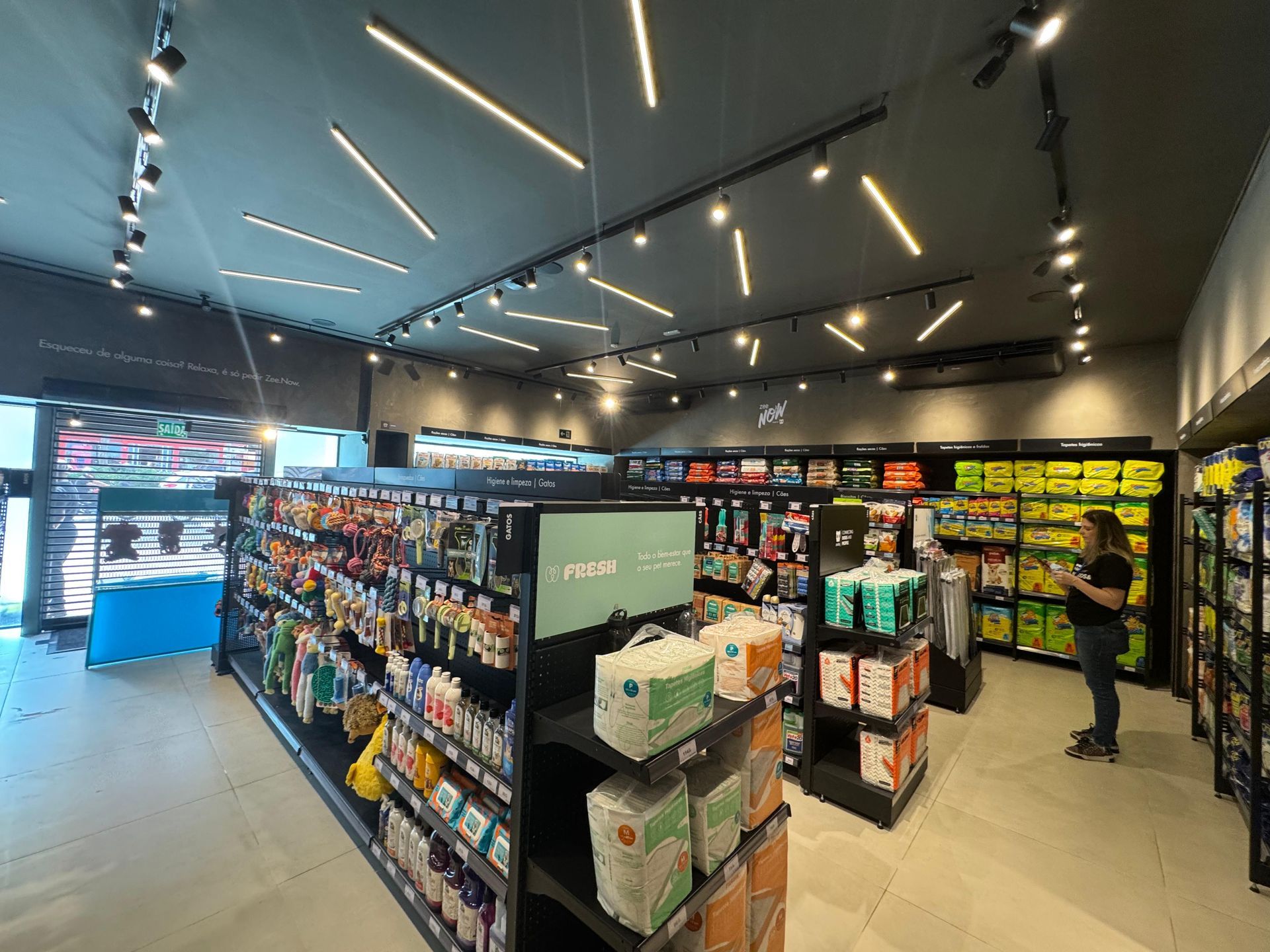 Corredor de loja de conveniência moderno e bem iluminado, com prateleiras repletas de salgadinhos e bebidas; uma pessoa está parada perto do fundo.