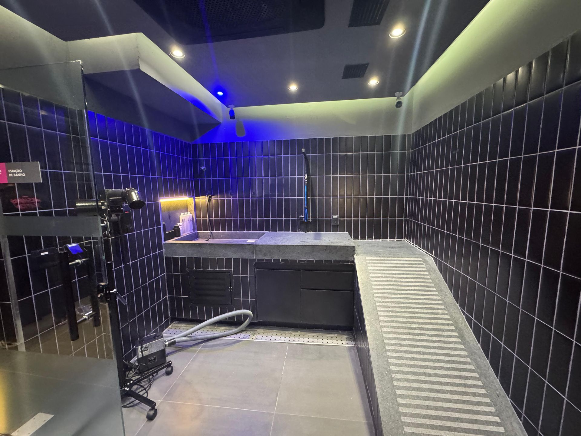 Sala vazia com azulejos pretos, plataforma elevada, iluminação azul e uma longa rampa metálica.