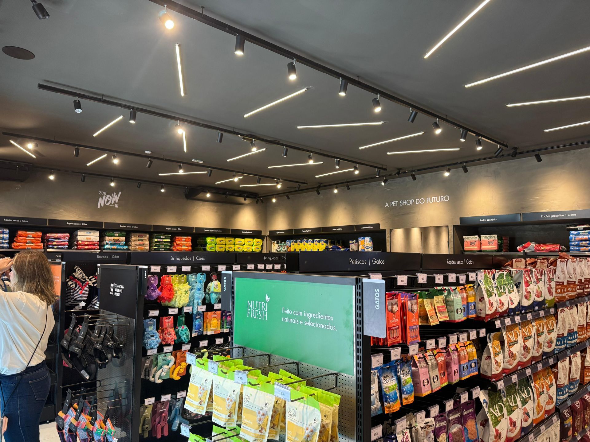 Corredor de supermercado com prateleiras de salgadinhos e bebidas embaladas sob luzes brilhantes no teto.