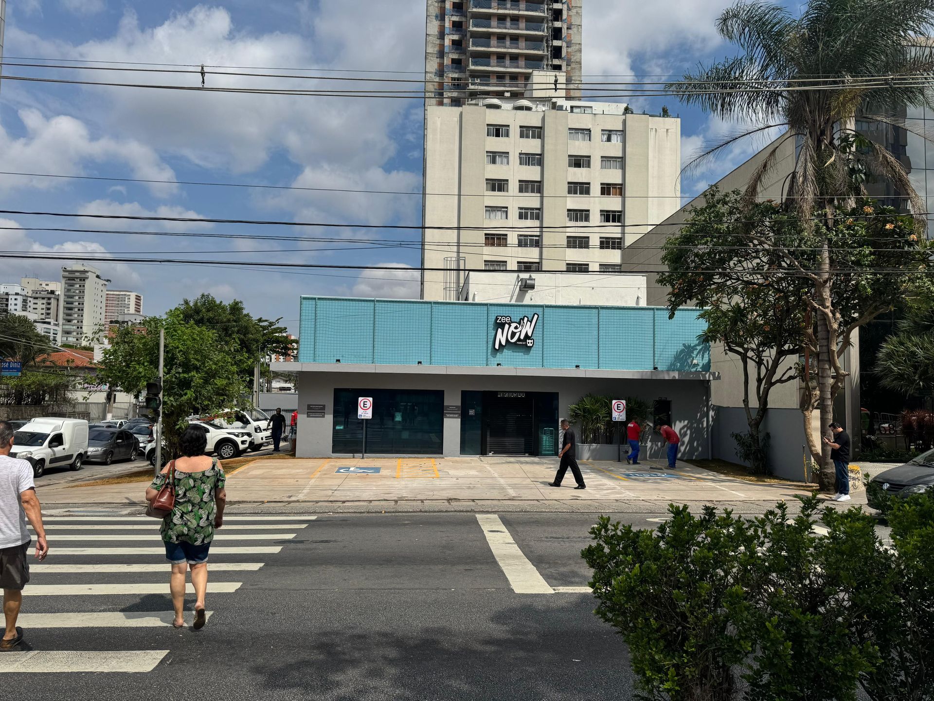 Vista da rua mostrando a fachada azul de uma loja perto de uma faixa de pedestres, com pedestres e prédios altos ao fundo.