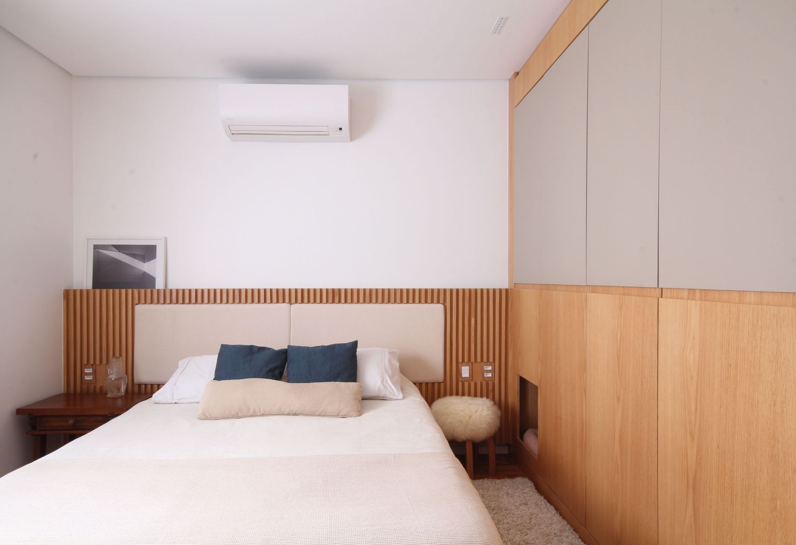 Quarto minimalista e luminoso com cama, detalhes em madeira, armários embutidos e ar condicionado.