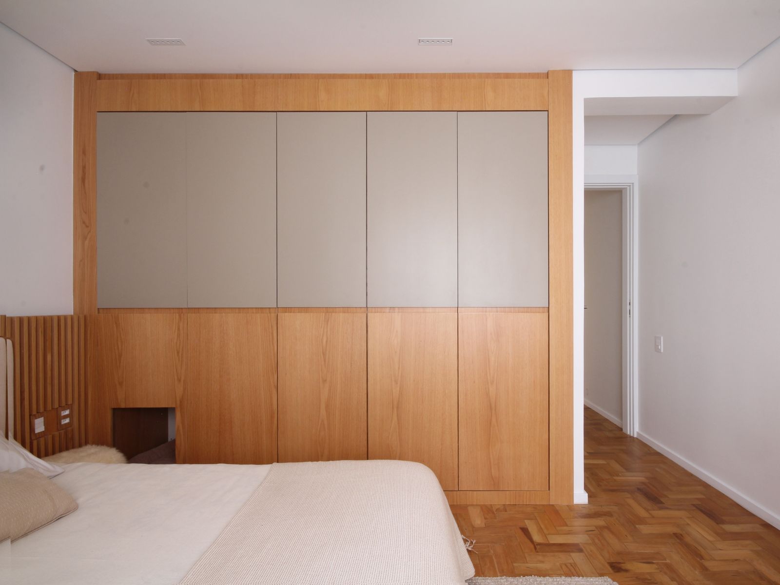 Quarto minimalista com armário embutido com painéis de madeira, paredes brancas e piso de madeira em espinha de peixe.