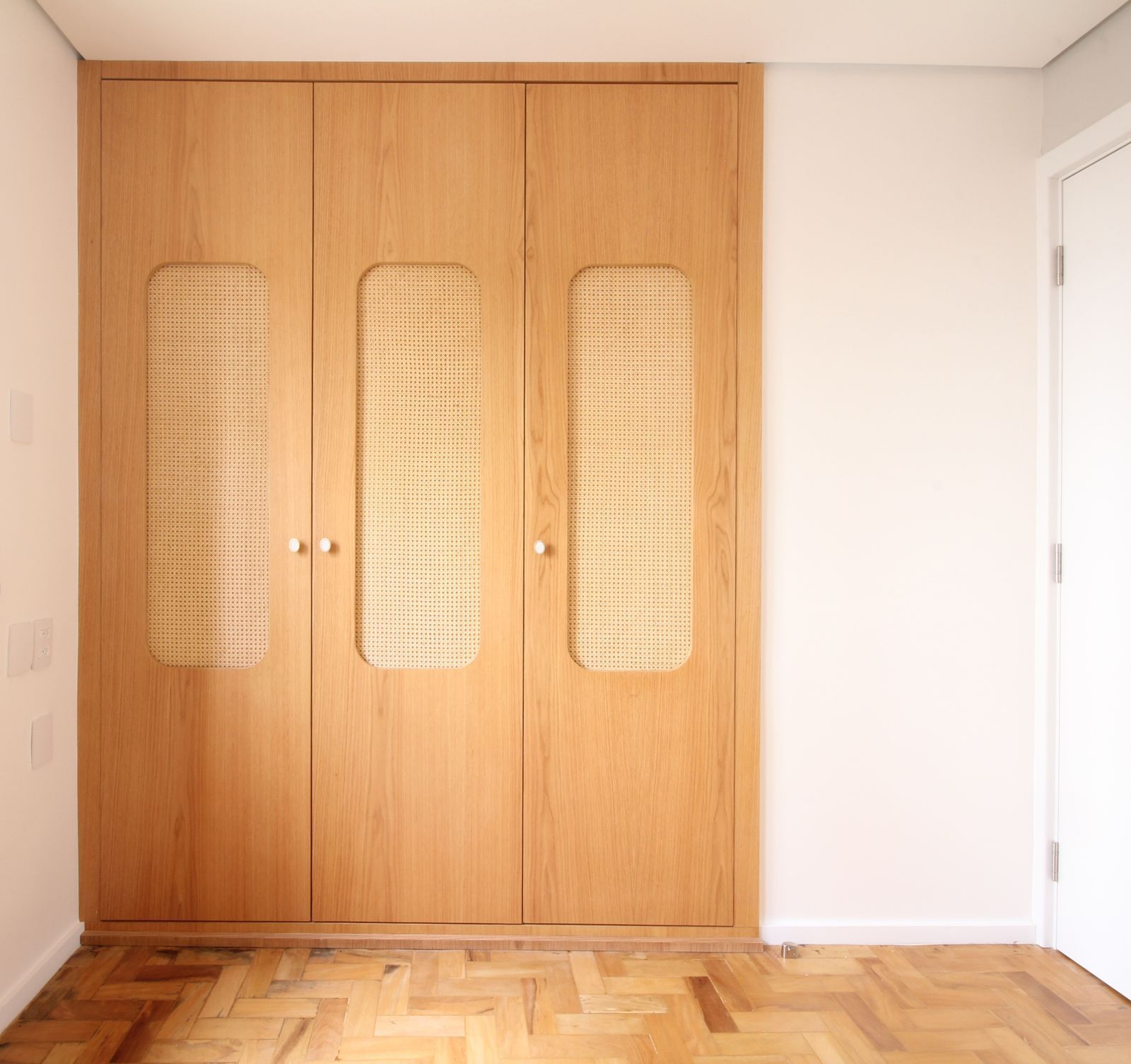 Guarda-roupa de madeira com painéis trançados em um quarto iluminado ao lado de uma porta branca.