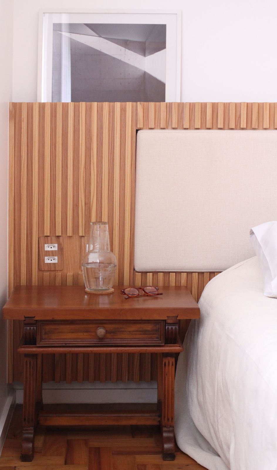 Quarto com mesa de cabeceira de madeira, abajur e cama branca ao lado de uma parede com detalhes em madeira listrada.