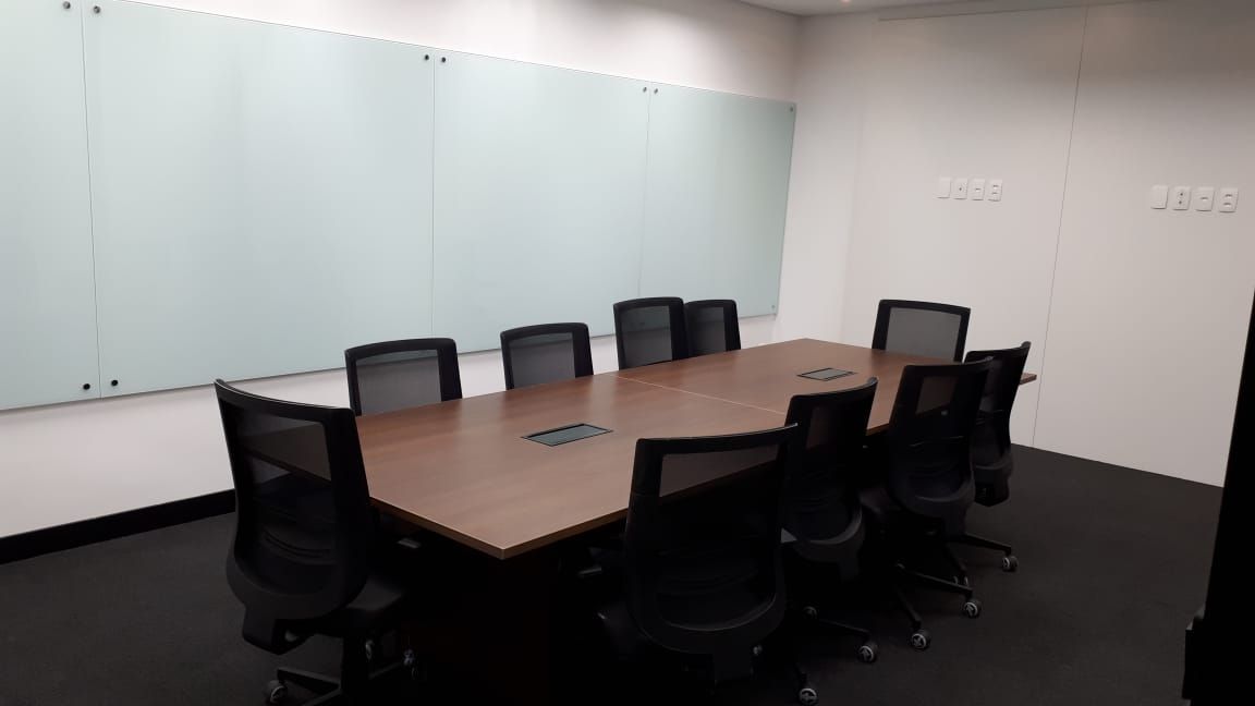 Sala de conferências vazia com uma longa mesa de madeira, cadeiras de escritório pretas e uma grande parede com quadro branco.