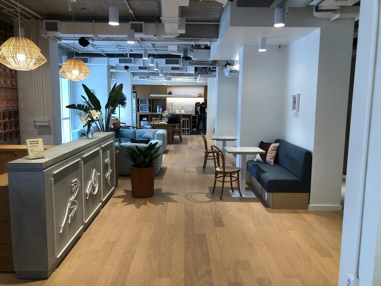Sala de estar moderna com piso de madeira, sofá cinza, balcão de bar, plantas e espaço de trabalho aberto ao fundo.
