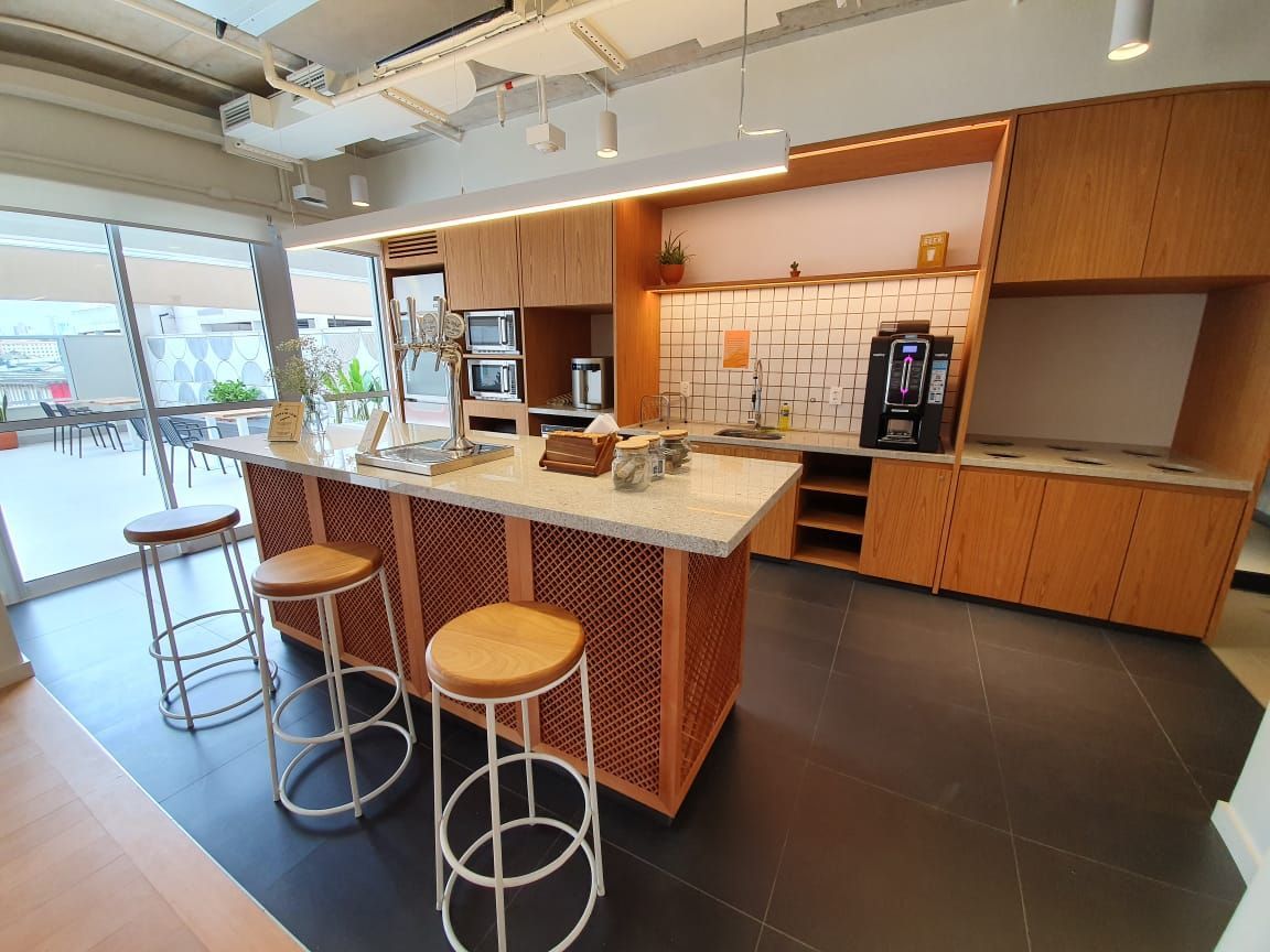 Cozinha de escritório moderna com ilha central, armários de madeira e espaço para café sob janelas amplas.