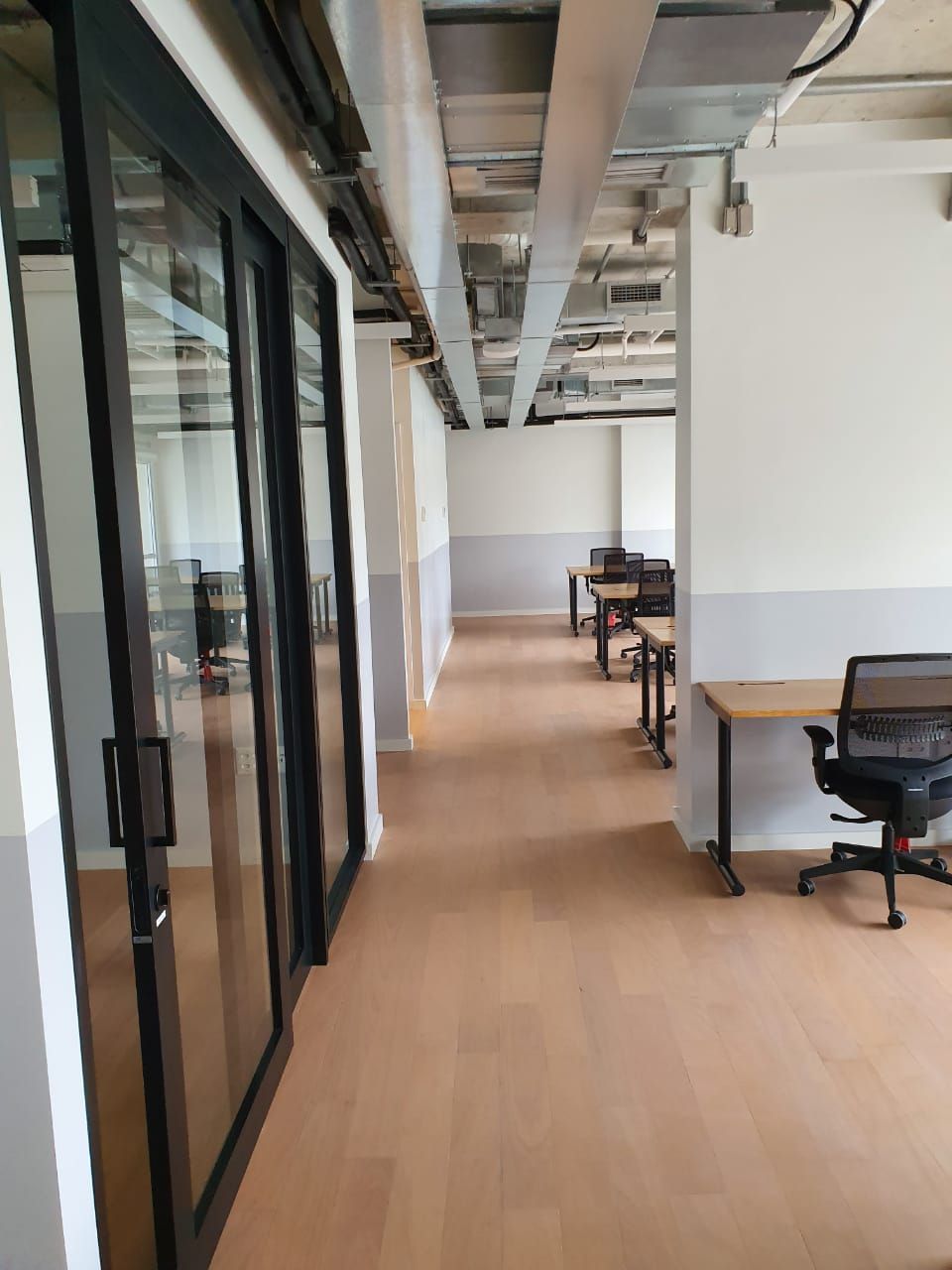Corredor de escritório moderno com paredes de vidro, mesas e cadeiras sobre piso de madeira clara.