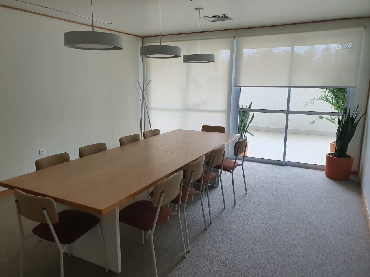 Sala de conferências moderna com uma longa mesa de madeira, cadeiras e grandes janelas com vasos de plantas.
