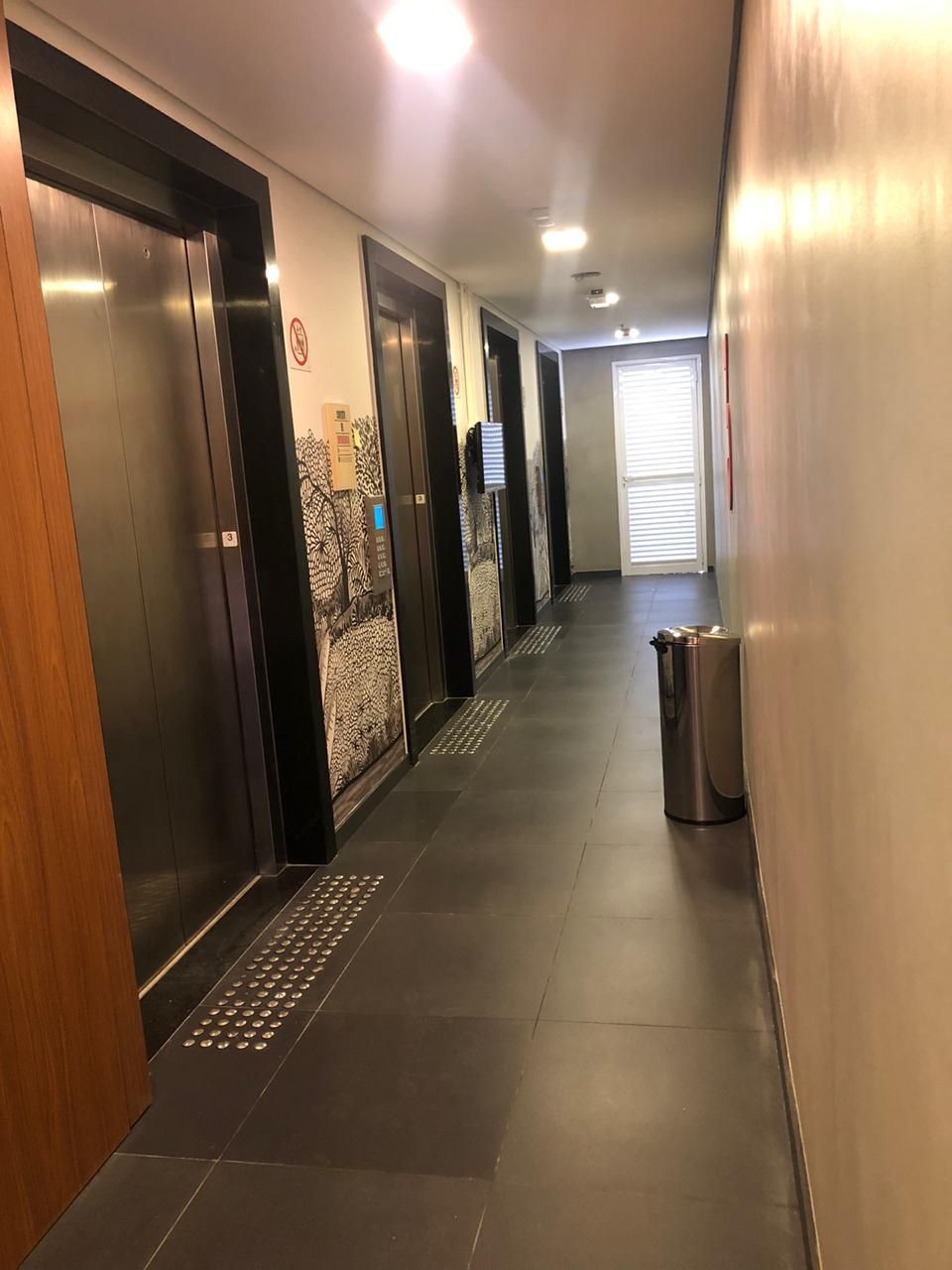 Corredor interno estreito com portas de elevador, piso de azulejo escuro e uma lixeira perto do final.