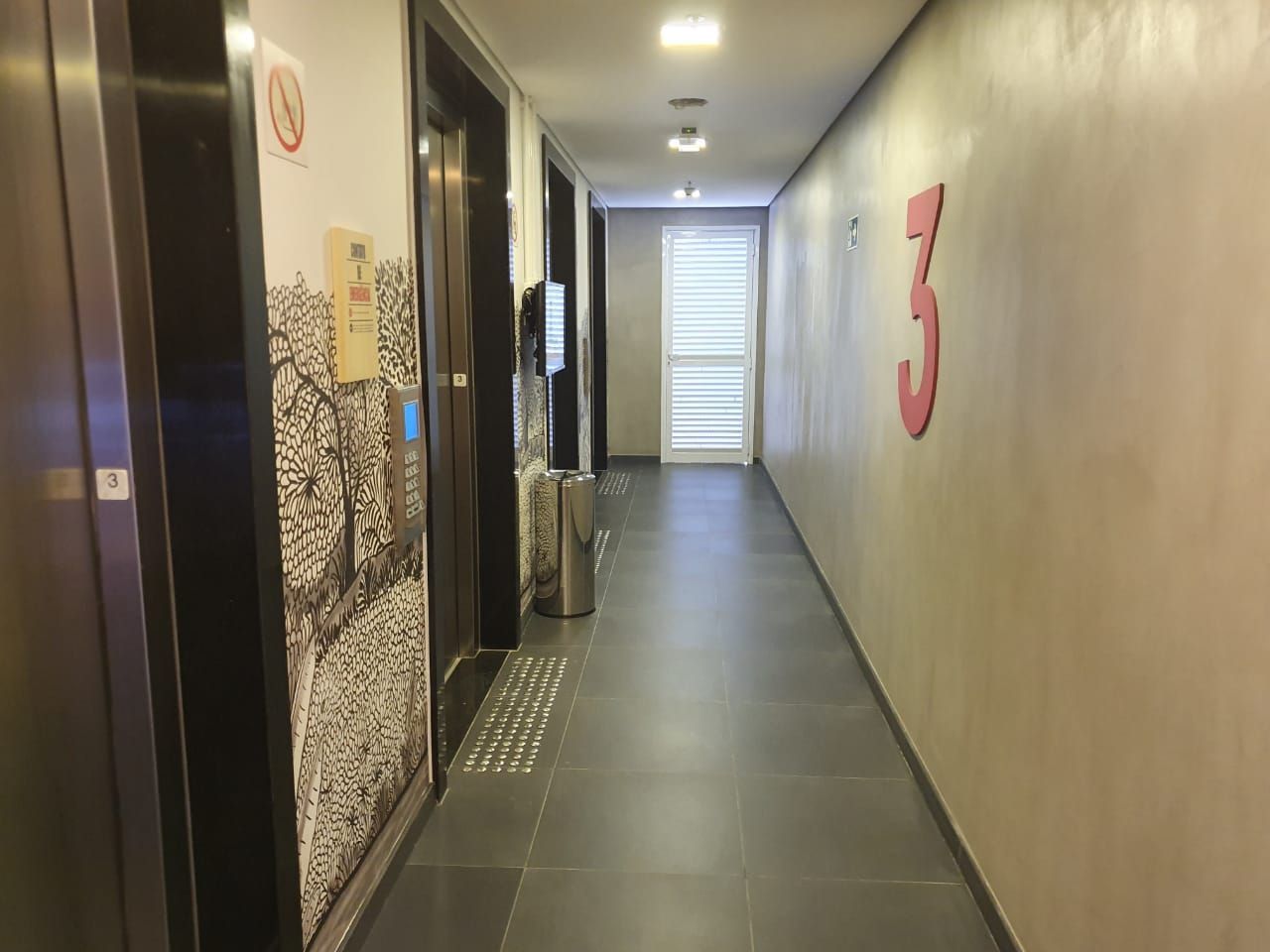 Corredor estreito de apartamento com portas de elevador, um grande número 3 vermelho na parede e uma luz forte no fundo.