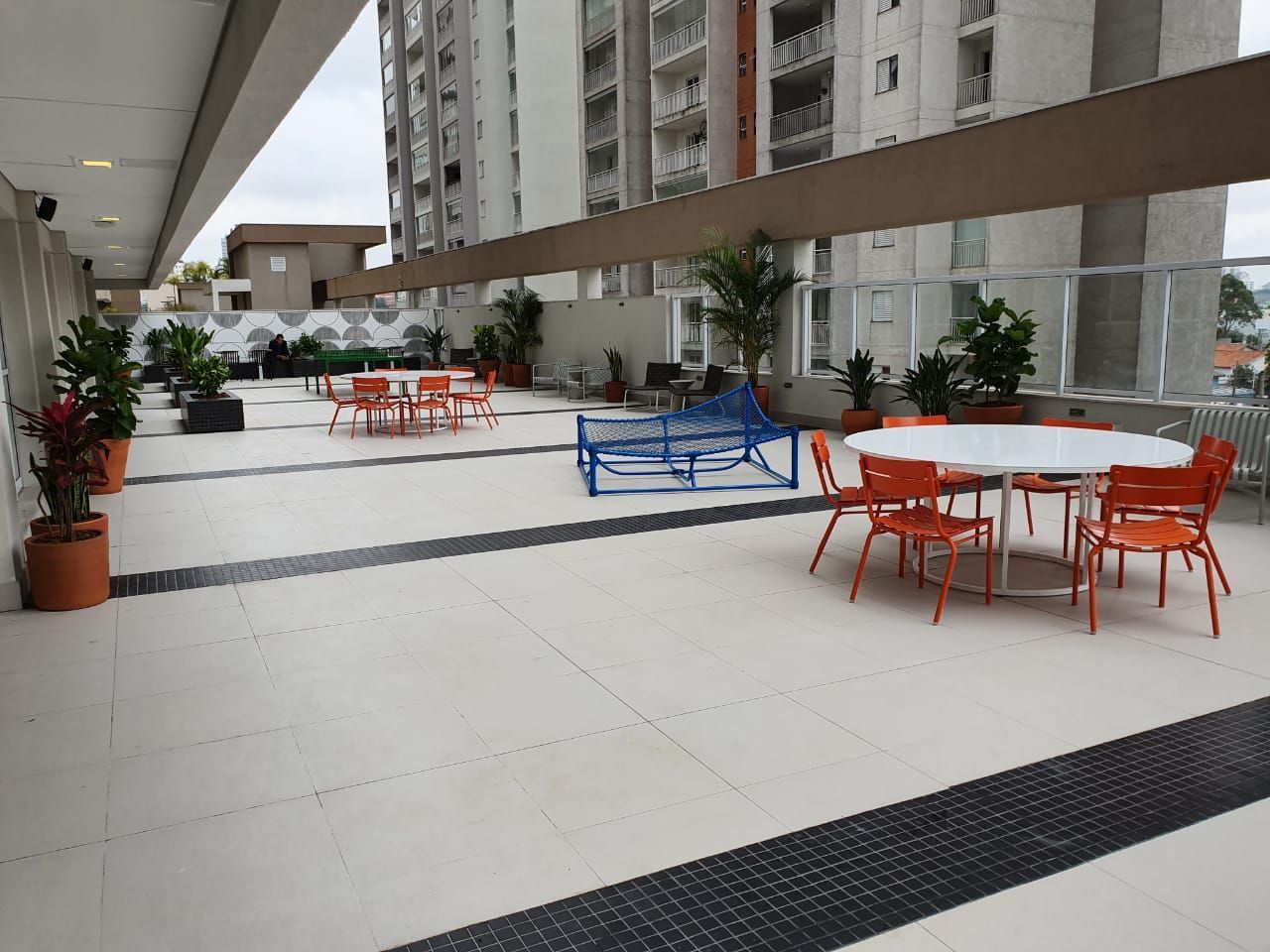 Terraço externo de hotel com cadeiras e mesas vermelhas e azuis ao lado de prédios altos de apartamentos.