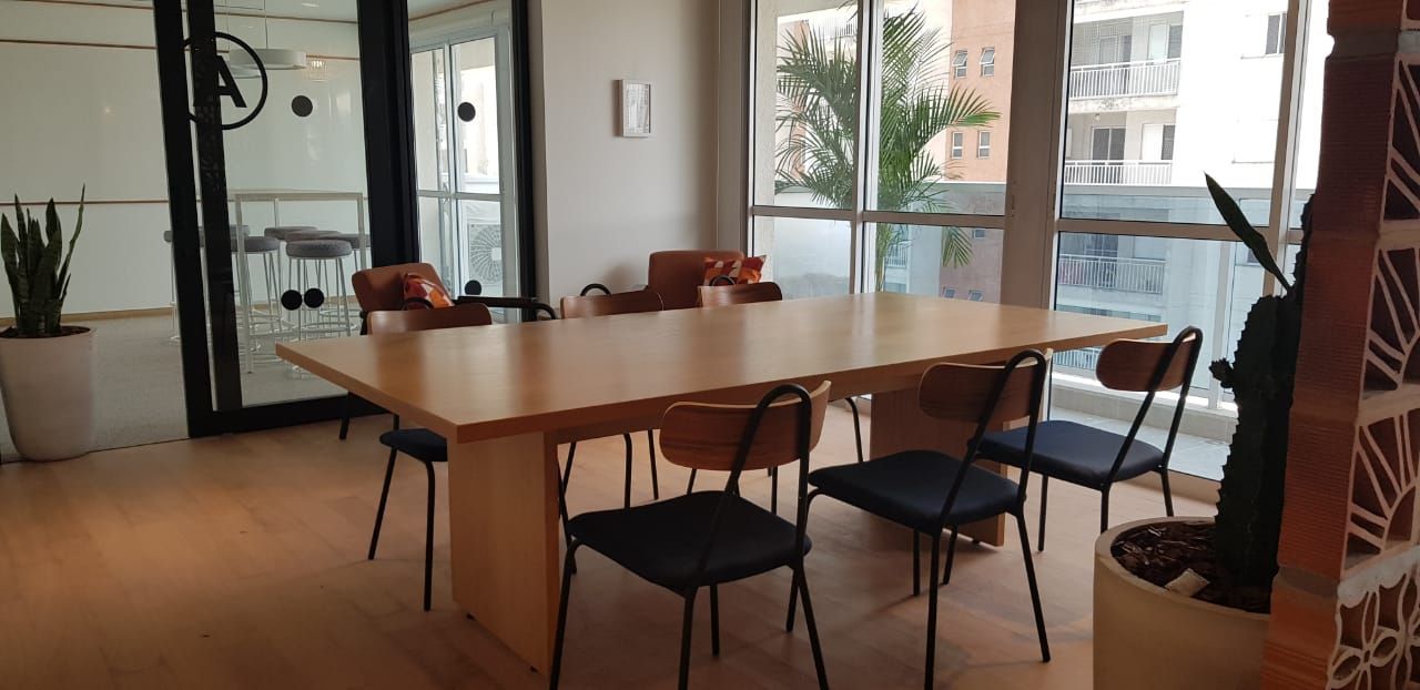 Sala de reuniões vazia com uma longa mesa de madeira, cadeiras e grandes janelas com vista para um corredor iluminado.