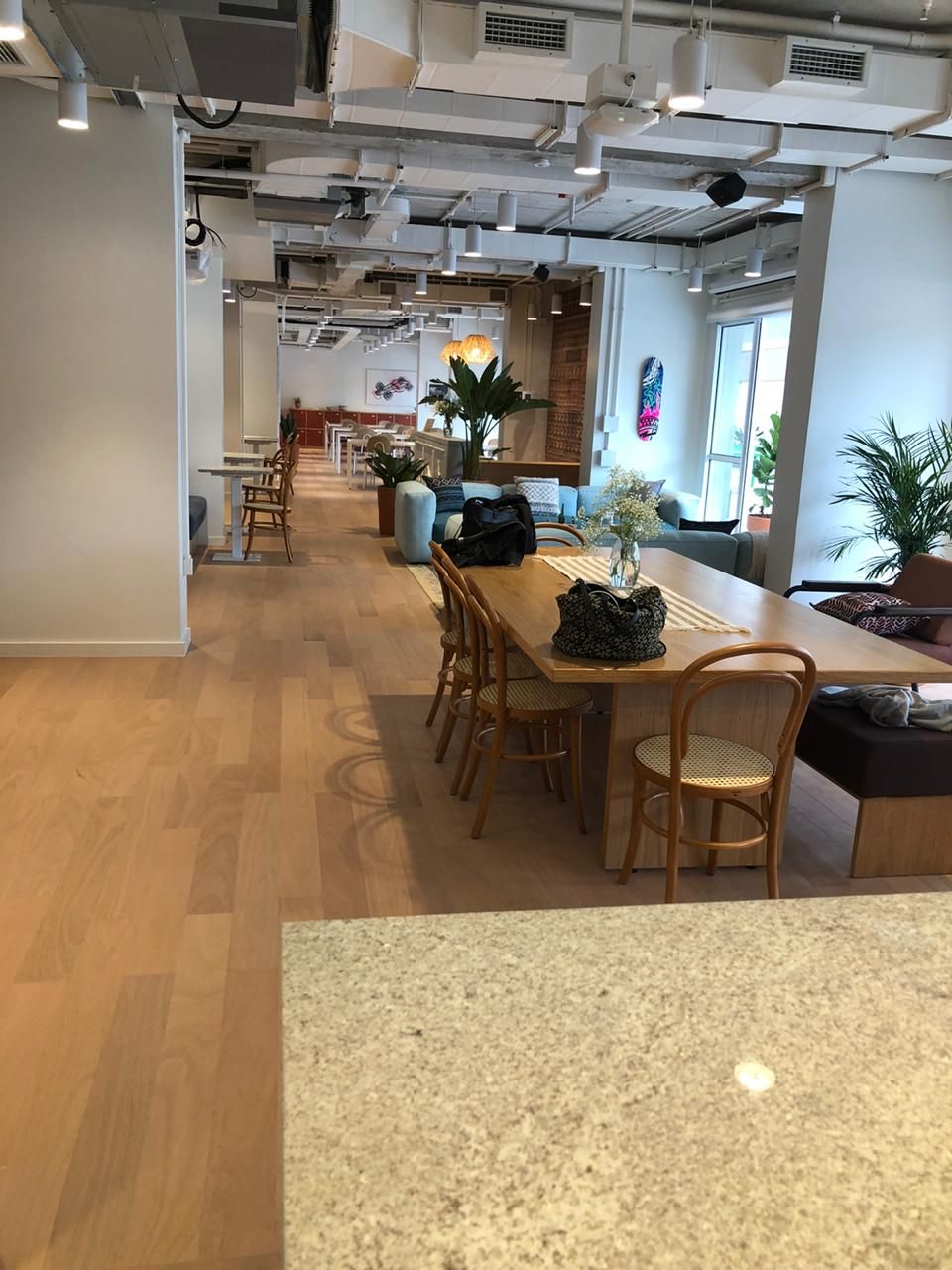 Café ou lounge de escritório moderno e luminoso, com mesas e cadeiras de madeira, plantas e teto aparente.