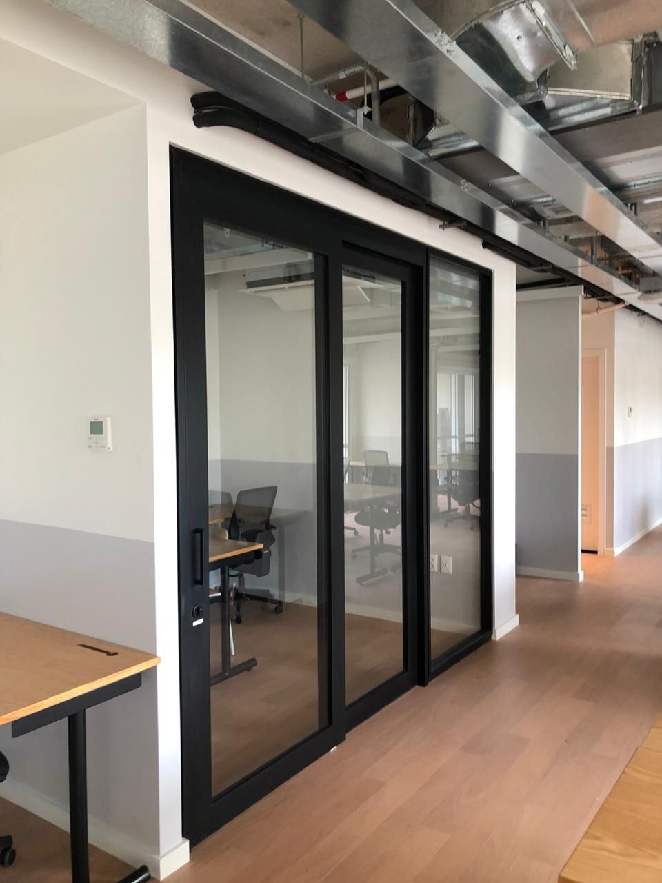 Corredor de escritório moderno com portas de sala de conferência em vidro com moldura preta e dutos de ar expostos no teto.