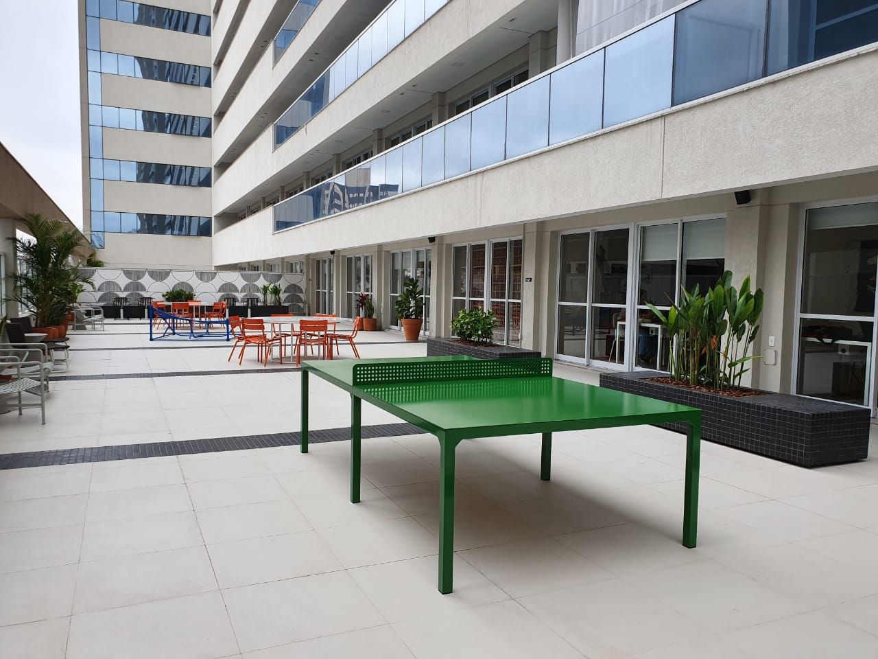 Pátio externo com mesa verde e cadeiras laranja ao lado de um prédio de apartamentos moderno.